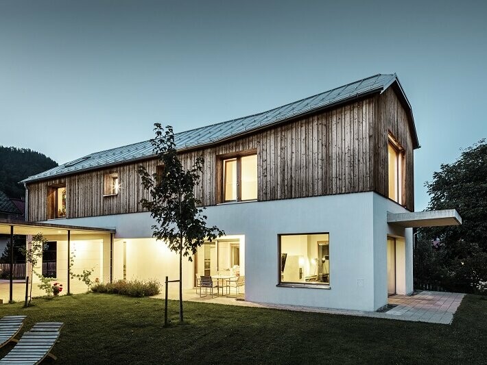 Frontalansicht des Einfamlienhauses im Ort Pruggern bei Dämmerung. Im Haus brennt Licht. Das Dach wurde mit PREFALZ in der Farbe Naturblank eingedeckt.