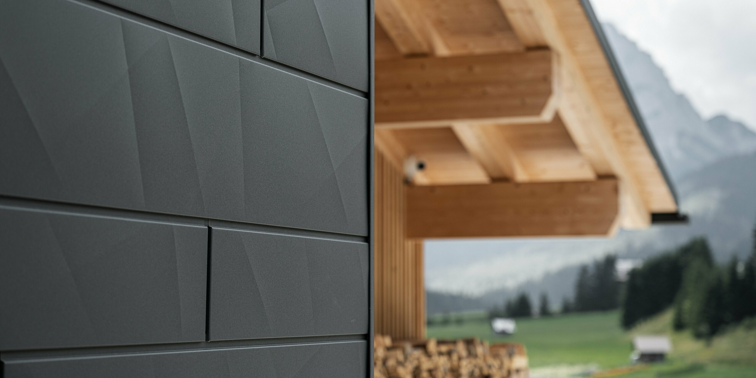 Close-up van de Siding.X gevelpanelen in P.10 zwart van PREFA aan de Casa Hoffe in Sappada, Italië. Het gevelsysteem van aluminium is vakkundig aangebracht in een modern, abstract ontwerp dat een aantrekkelijk contrast vormt met de zichtbare houten elementen van het dakgebinte. De innovatieve geveloplossing past naadloos in de alpine omgeving en staat voor de combinatie van functionaliteit, duurzaamheid en esthetische bouwcultuur.