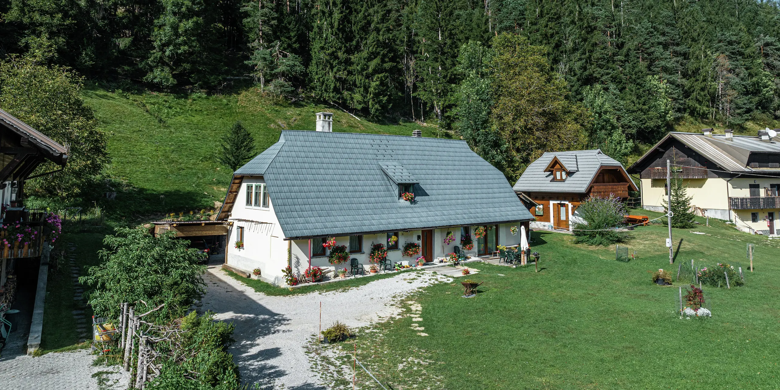 Luftaufnahme eines idyllischen Einfamilienhauses in Tarvis, Italien, das seit 1972 von langlebigen PREFA Dachplatten geschützt wird. Das großflächige, witterungsbeständige Aluminiumdach fügt sich harmonisch in die grüne Alpenlandschaft ein. Die liebevoll mit Blumen geschmückte Fassade und die gemütliche Terrasse verleihen dem Haus eine einladende Atmosphäre. Umgeben von traditionellen Häusern mit Holzfassaden und dichtem Nadelwald bietet das Anwesen eine perfekte Mischung aus Naturverbundenheit und architektonischer Beständigkeit.