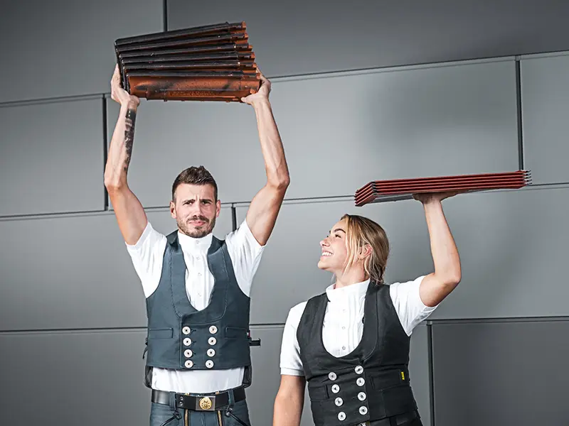 Op de foto zijn twee personen te zien die elk een stapel dakbedekkingsmateriaal in hun handen houden. De persoon aan de linkerkant houdt een stapel dakpannen boven zijn hoofd, terwijl de persoon aan de rechterkant een stapel aluminium dakplaten ook boven zijn hoofd houdt. Beiden dragen een traditionele ambachtsman-outfit met een vest en een wit overhemd. De achtergrond is modern en minimalistisch, met grijze muren. Beide personen zien er geconcentreerd uit en glimlachen.