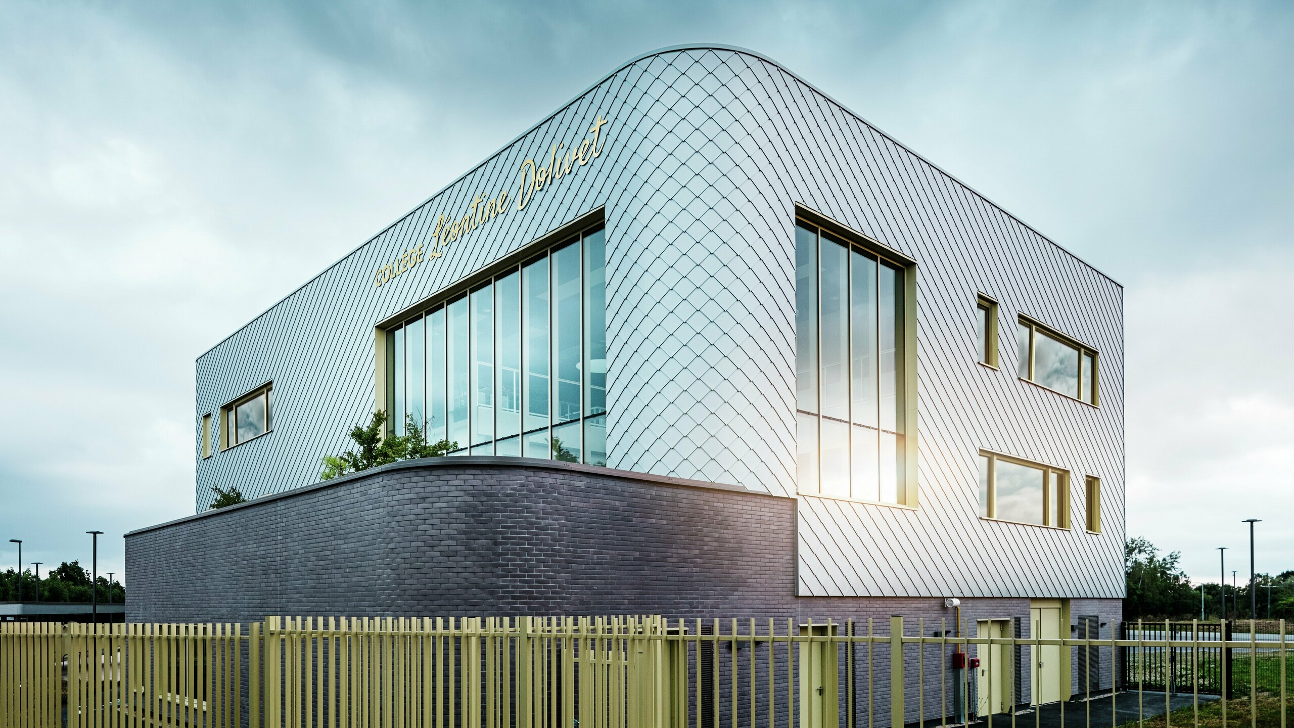 Das Foto zeigt die markante Ecke des Collège Léontine Dolivet mit geschwungener Aluminiumfassade aus silbermetallic PREFA Wandrauten. Große Glasflächen reflektieren das Licht, während gelbe Fensterrahmen Akzente setzen. Im Vordergrund verläuft ein heller Zaun entlang des Gebäudes, der auf einem dunklen Sockel aus Ziegeln steht. Der Himmel ist bewölkt.