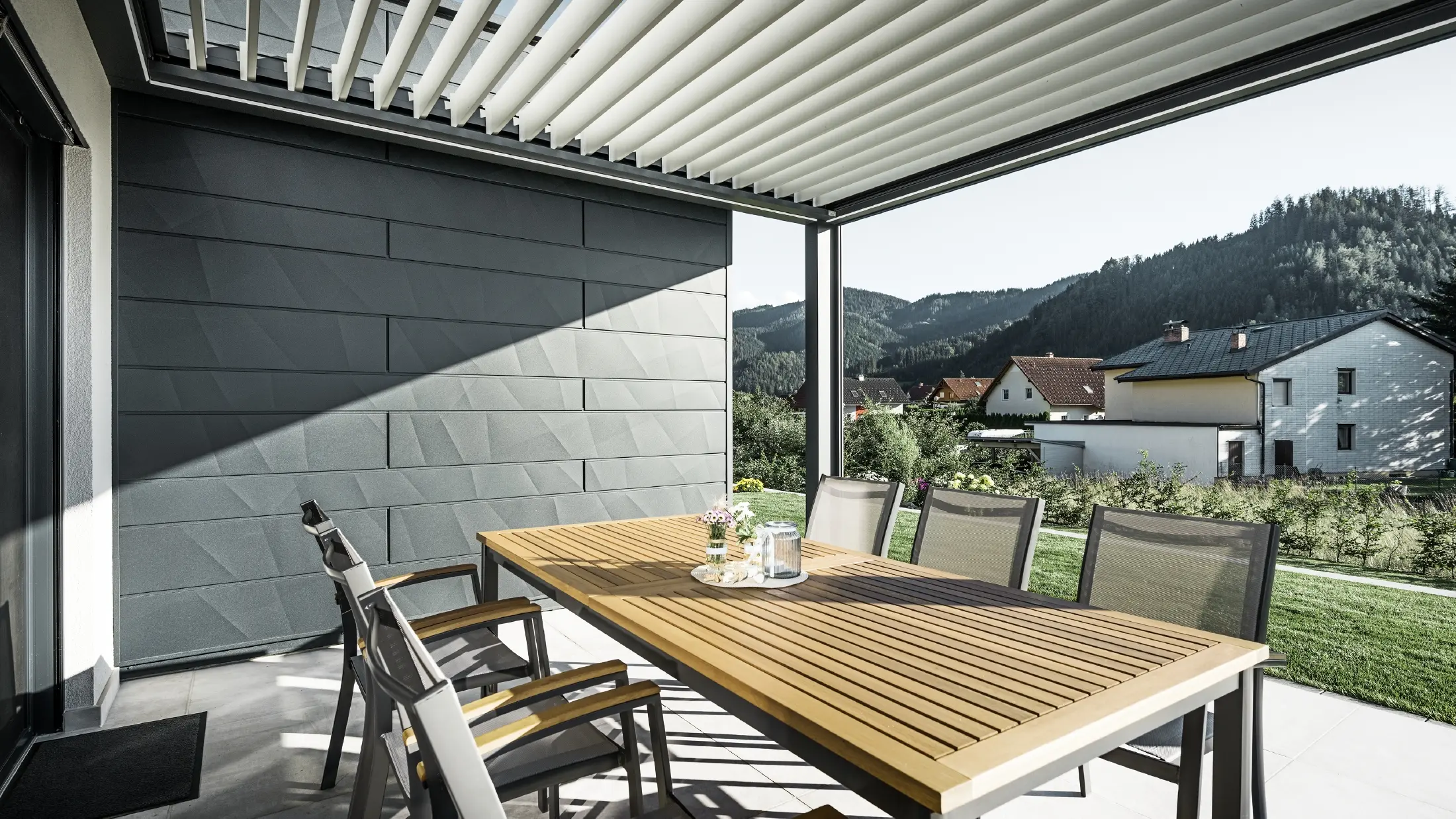 Met lamellen overdekt, beschut terras met tafel en stoelen, een gevelkant is bekleed met de PREFA Siding.X gevelpanelen in antraciet.