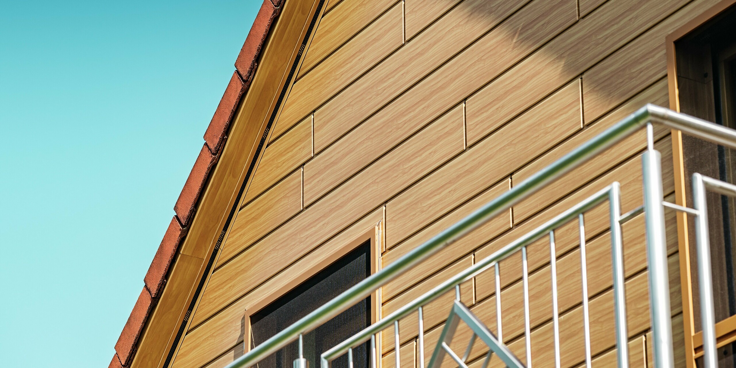 Detailaufnahme des sanierten Giebels eines Einfamilienhauses in Blieskastel. Der Giebel wurde mit PREFA Sidings in der Farbe Eiche Natur verkleidet, wodurch er eine warme, moderne Holzoptik erhält. Die feine Struktur und der natürliche Farbton der Aluminiumfassade sehen aus wie echtes Holz und setzen einen starken visuellen Akzent unter dem klaren Sommerhimmel. Gleichzeitig bleibt die Oberfläche dauerhaft witterungsbeständig, farbstabil und nahezu wartungsfrei. Sie ist ein langlebiges Highlight, das sich harmonisch in die Balkongeländer und das rote Ziegeldach einfügt.