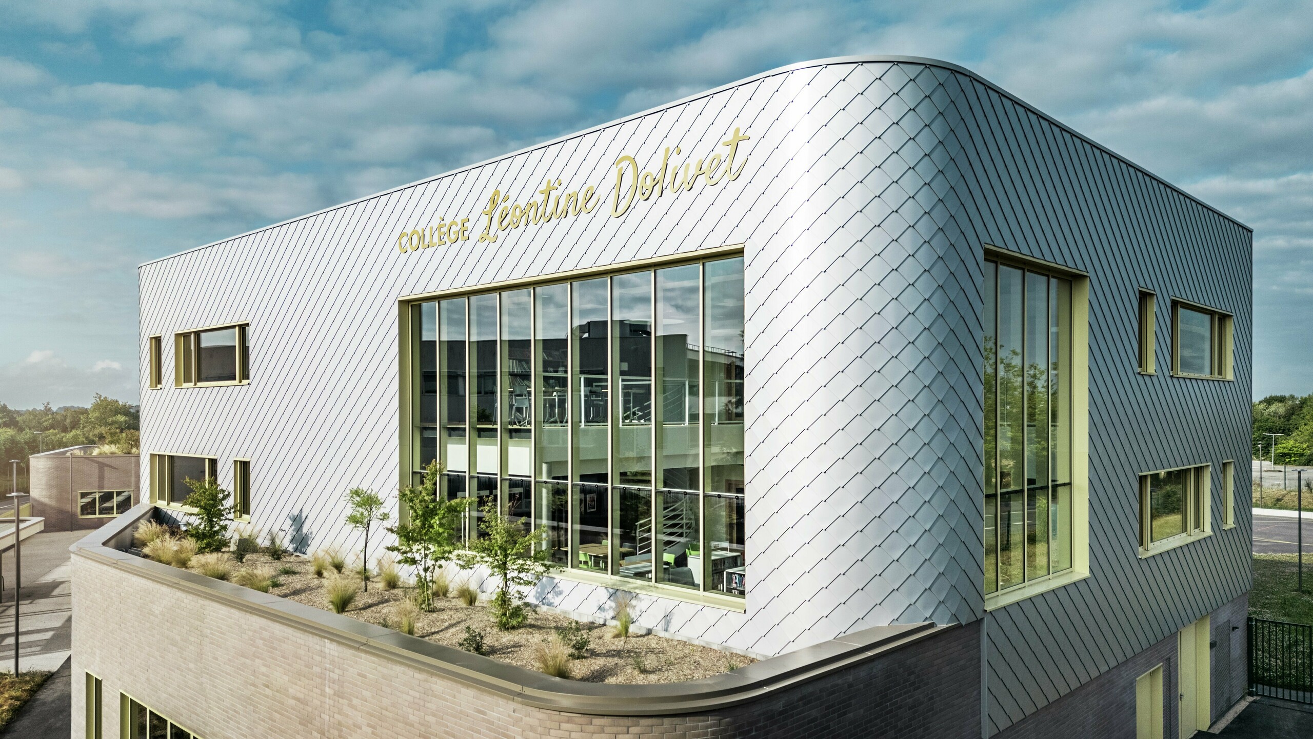 Das Foto zeigt eine markante Gebäudeecke des Collège Léontine Dolivet in Cesson-Sévigné. Die Fassade besteht aus silbermetallic PREFA Wandrauten, die in einem diagonalen Muster angeordnet sind und das Licht reflektieren. Auffällig ist die geschwungene Form der oberen Etage, die dem Bau eine weiche, moderne Anmutung verleiht. Große Glasfronten öffnen den Blick ins Innere, während gelbe Fensterrahmen lebendige Akzente setzen. Im Vordergrund befindet sich ein bepflanzter Bereich mit Kies und Sträuchern, der die Architektur harmonisch ergänzt. Der Himmel ist leicht bewölkt, was die metallische Oberfläche betont.