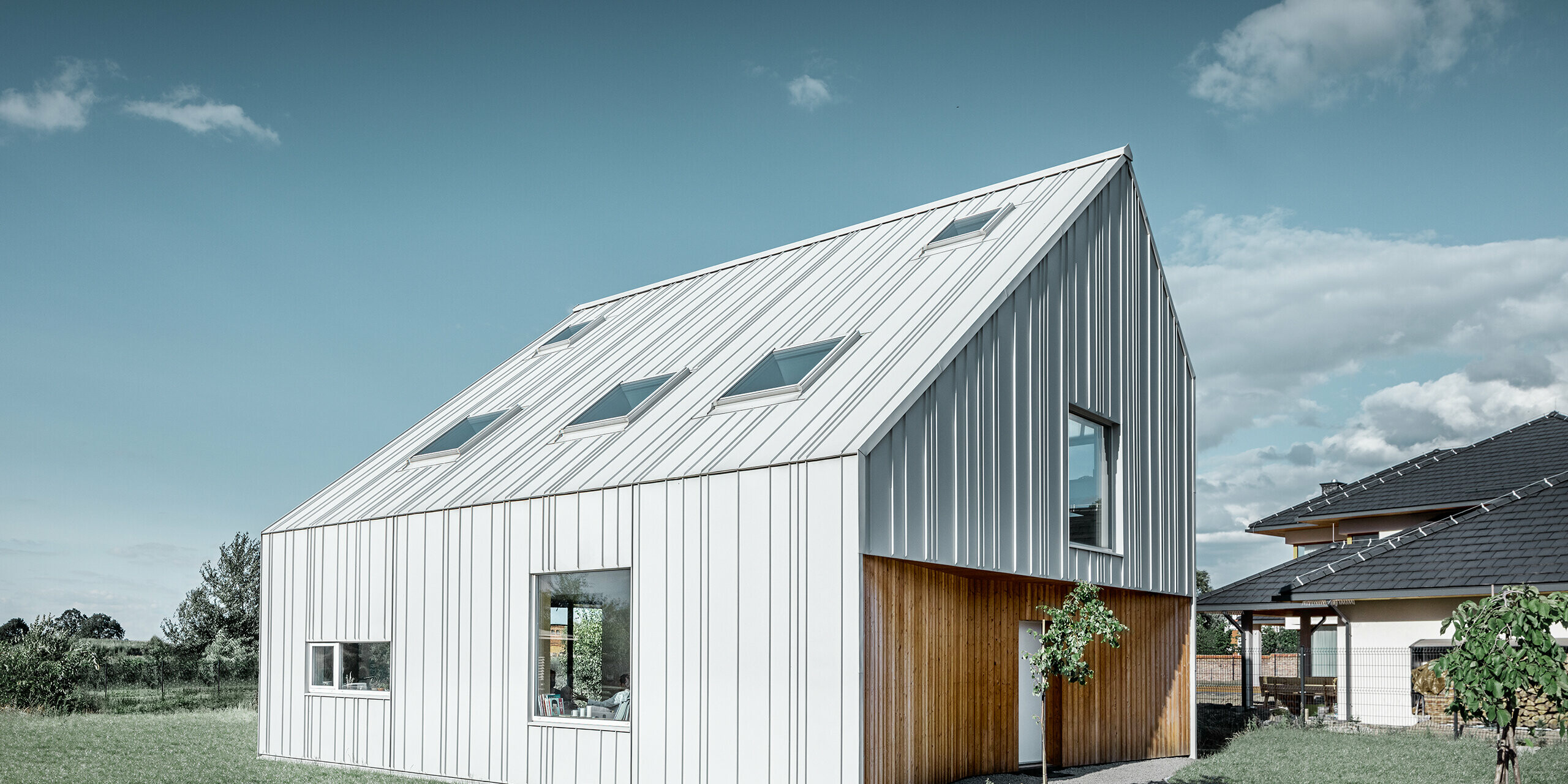 Modern vormgegeven eengezinswoning met zadeldak, de volledige buitenkant is bekleed met PREFALZ in prefawit. Daarbij lopen de verschillend brede panelen van het dak door over de gevel. In het dak bevinden zich verschillende dakvensters.