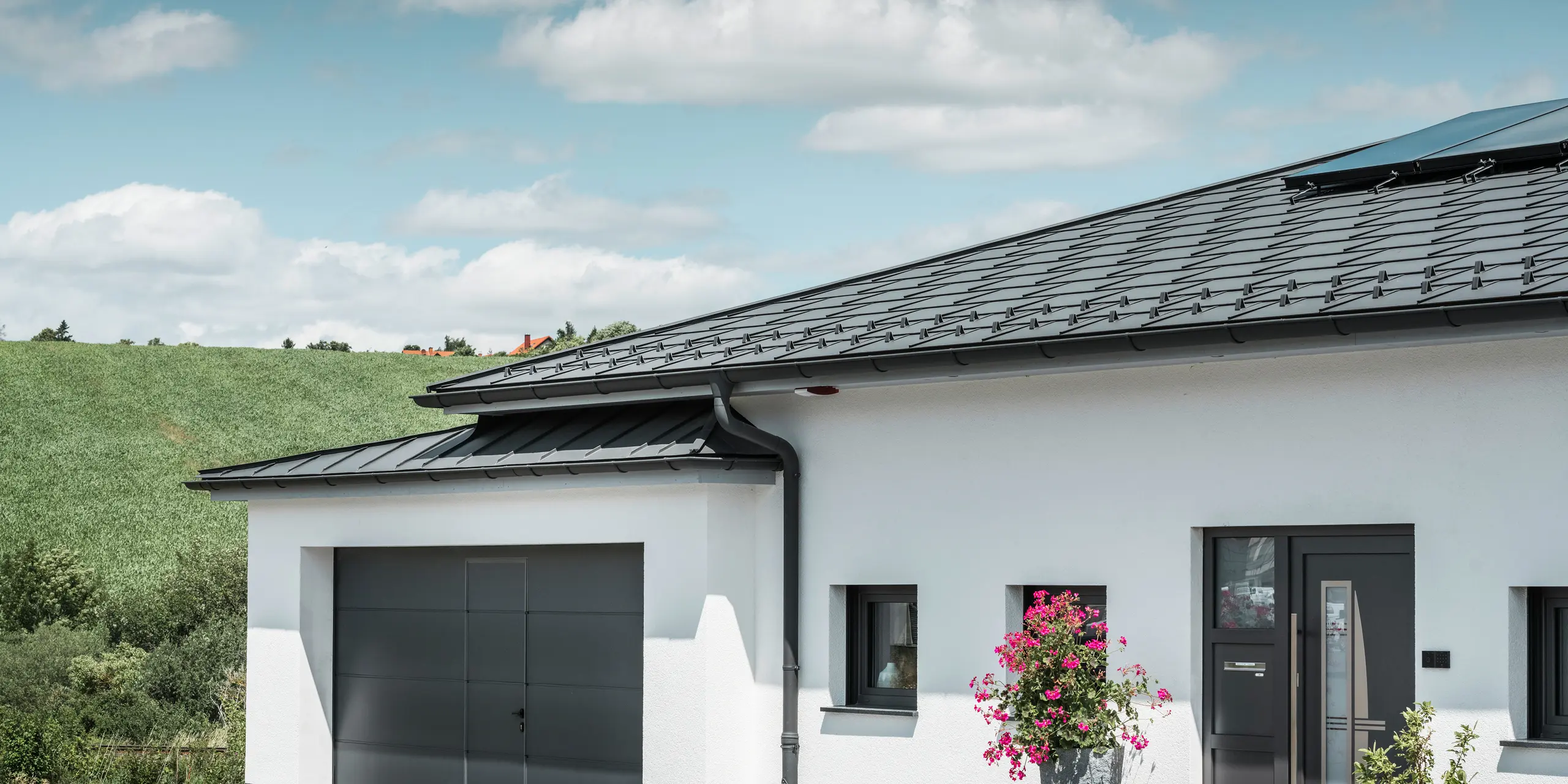 De foto toont de voorkant van een eengezinswoning in Ilmenau, Thüringen, met een elegant PREFA-dak in P.10 donkergrijs. De dakplaat R.16 geeft het huis een moderne uitstraling, terwijl het geïntegreerde PREFA dakafvoersysteem met zijn donkergrijze hanggoot en afvoerpijp zorgt voor een efficiënte afvoer van regenwater. De garage, uitgevoerd in donkergrijs PREFALZ, past harmonieus in het totaalbeeld en onderstreept de kwaliteit en stijl van PREFA aluminiumproducten.