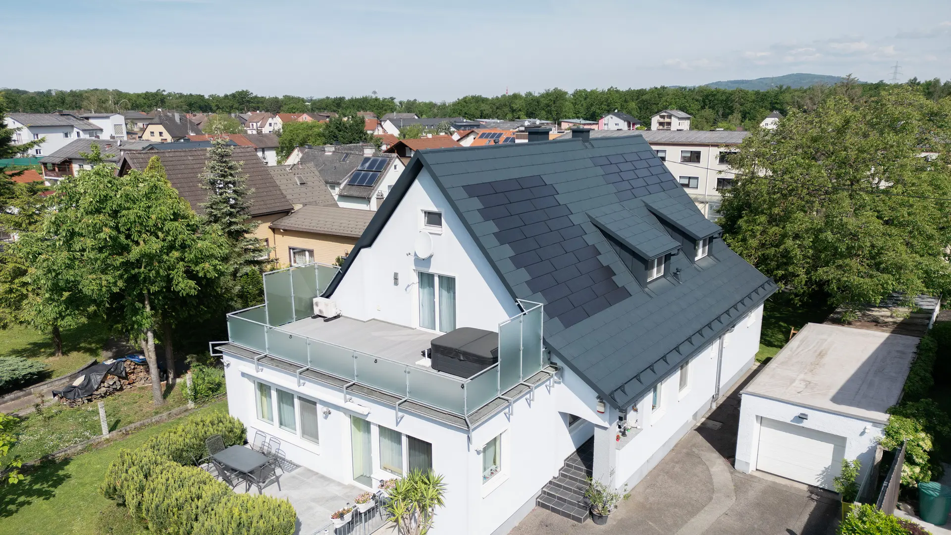 Vogelvluchtperspectief op een eengezinswoning na de dakrenovatie. Het dak is nu bedekt met grote PREFA solar-dakpannen en de PREFA-dakpan R.16 in P.10 antraciet. De solar-dakpannen zijn gelijkmatig verdeeld en zorgen voor een moderne, duurzame oplossing. Het gebouw ziet er nu modern en milieuvriendelijk uit. Het balkon op de voorgrond heeft een glazen balustrade en bevindt zich op de bovenste verdieping van het gebouw.