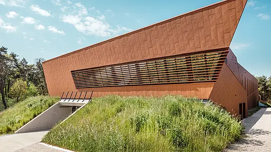 Das auffällige Trinkwasserpumpwerk WML in Lottum verfügt über eine expressiv geneigte Gebäudespitze und eine dynamisch geformte PREFA Siding.X-Aluminiumfassade in warmer Rostoptik. Die facettierte Oberfläche erzeugt ein lebendiges Spiel aus Licht und Schatten, während die horizontalen Lamellenbänder architektonische Tiefe schaffen. Eingebettet in eine naturnahe Böschung wirkt das Gebäude wie eine skulpturale Landmarke. Die langlebigen, leichten und recycelbaren PREFA-Aluminium-Paneele überzeugen durch hohe Witterungsbeständigkeit und Formstabilität sowie eine nachhaltige, wartungsarme Gebäudehülle.