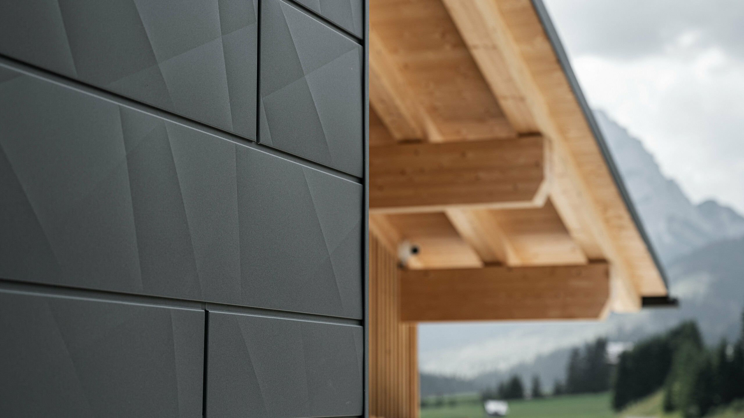 Close-up van de Siding.X gevelpanelen in P.10 zwart van PREFA aan de Casa Hoffe in Sappada, Italië. Het gevelsysteem van aluminium is vakkundig aangebracht in een modern, abstract ontwerp dat een aantrekkelijk contrast vormt met de zichtbare houten elementen van het dakgebinte. De innovatieve geveloplossing past naadloos in de alpine omgeving en staat voor de combinatie van functionaliteit, duurzaamheid en esthetische bouwcultuur.