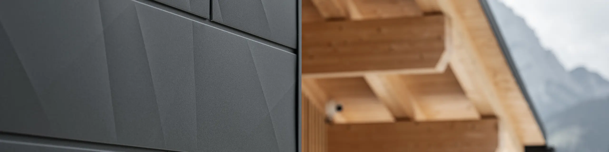 Detailfoto van de Siding.X gevelpanelen in P.10 zwart van PREFA aan Casa Hoffe in Sappada, Italië. Het gevelsysteem van aluminium is vakkundig aangebracht in een modern, abstract design, dat een aantrekkelijk contract vormt met de zichtbare houten elementen van de dakconstructie. De innovatieve geveloplossing past perfect in de alpiene omgeving en staat symbool voor de verbinding van functionaliteit, bestendigheid en esthetische bouwcultuur.