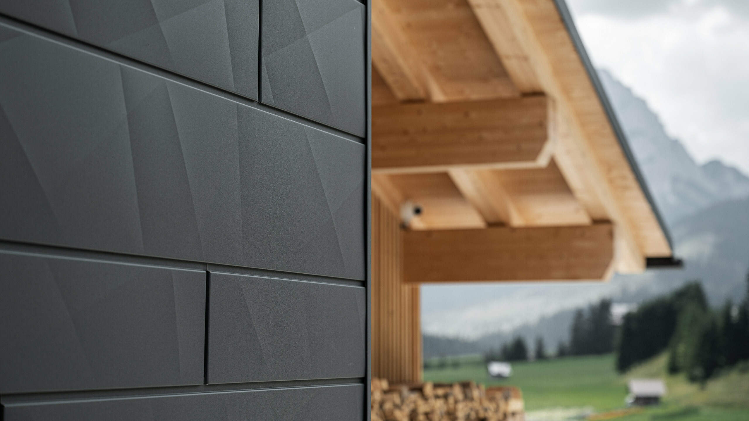 Close-up van de Siding.X gevelpanelen in P.10 zwart van PREFA aan de Casa Hoffe in Sappada, Italië. Het gevelsysteem van aluminium is vakkundig aangebracht in een modern, abstract ontwerp dat een aantrekkelijk contrast vormt met de zichtbare houten elementen van het dakgebinte. De innovatieve geveloplossing past naadloos in de alpine omgeving en staat voor de combinatie van functionaliteit, duurzaamheid en esthetische bouwcultuur.