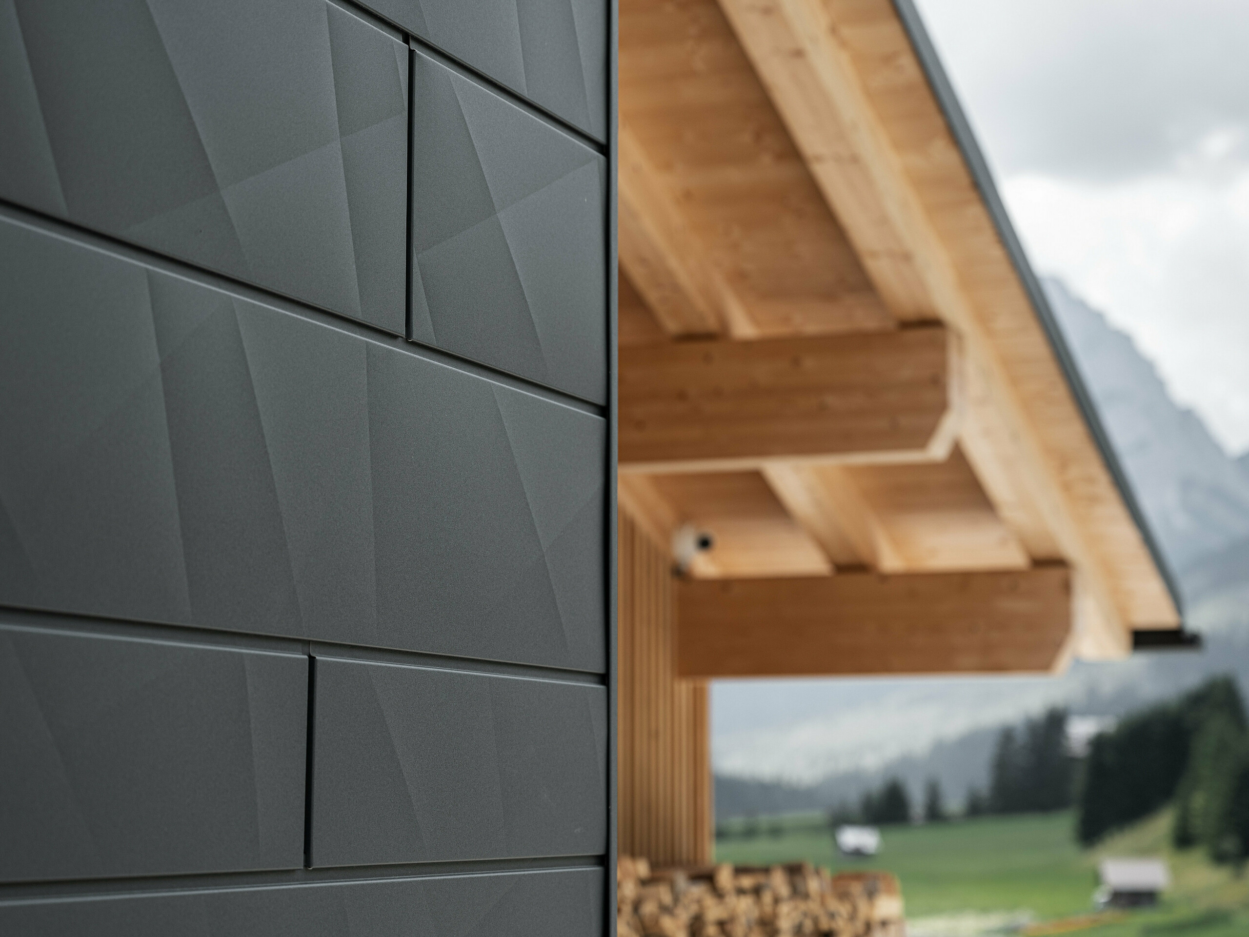 Detailfoto van de Siding.X gevelpanelen in P.10 zwart van PREFA aan Casa Hoffe in Sappada, Italië. Het gevelsysteem van aluminium is vakkundig aangebracht in een modern, abstract design, dat een aantrekkelijk contract vormt met de zichtbare houten elementen van de dakconstructie. De innovatieve geveloplossing past perfect in de alpiene omgeving en staat symbool voor de verbinding van functionaliteit, bestendigheid en esthetische bouwcultuur.