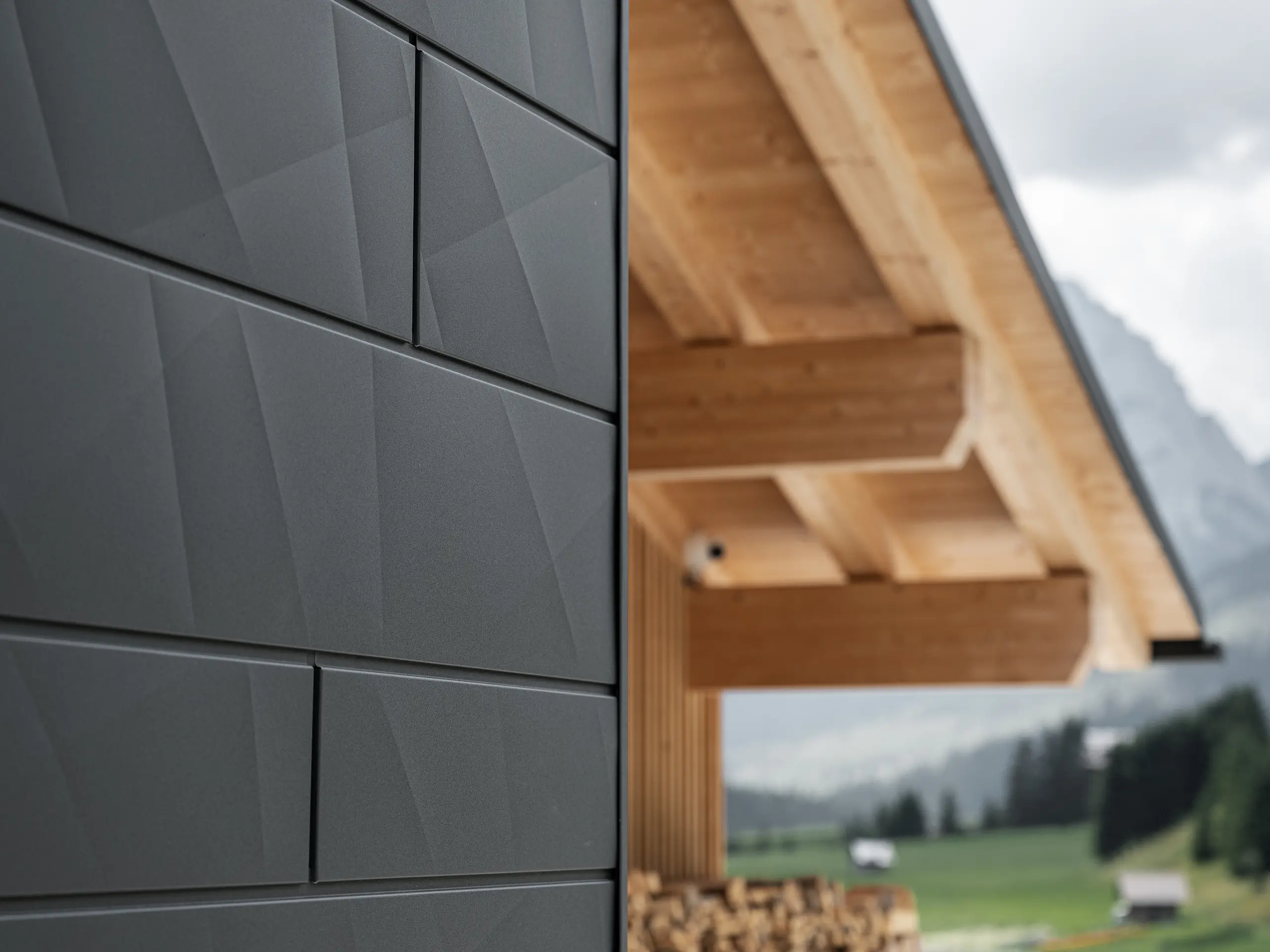 Detailfoto van de Siding.X gevelpanelen in P.10 zwart van PREFA aan Casa Hoffe in Sappada, Italië. Het gevelsysteem van aluminium is vakkundig aangebracht in een modern, abstract design, dat een aantrekkelijk contract vormt met de zichtbare houten elementen van de dakconstructie. De innovatieve geveloplossing past perfect in de alpiene omgeving en staat symbool voor de verbinding van functionaliteit, bestendigheid en esthetische bouwcultuur.