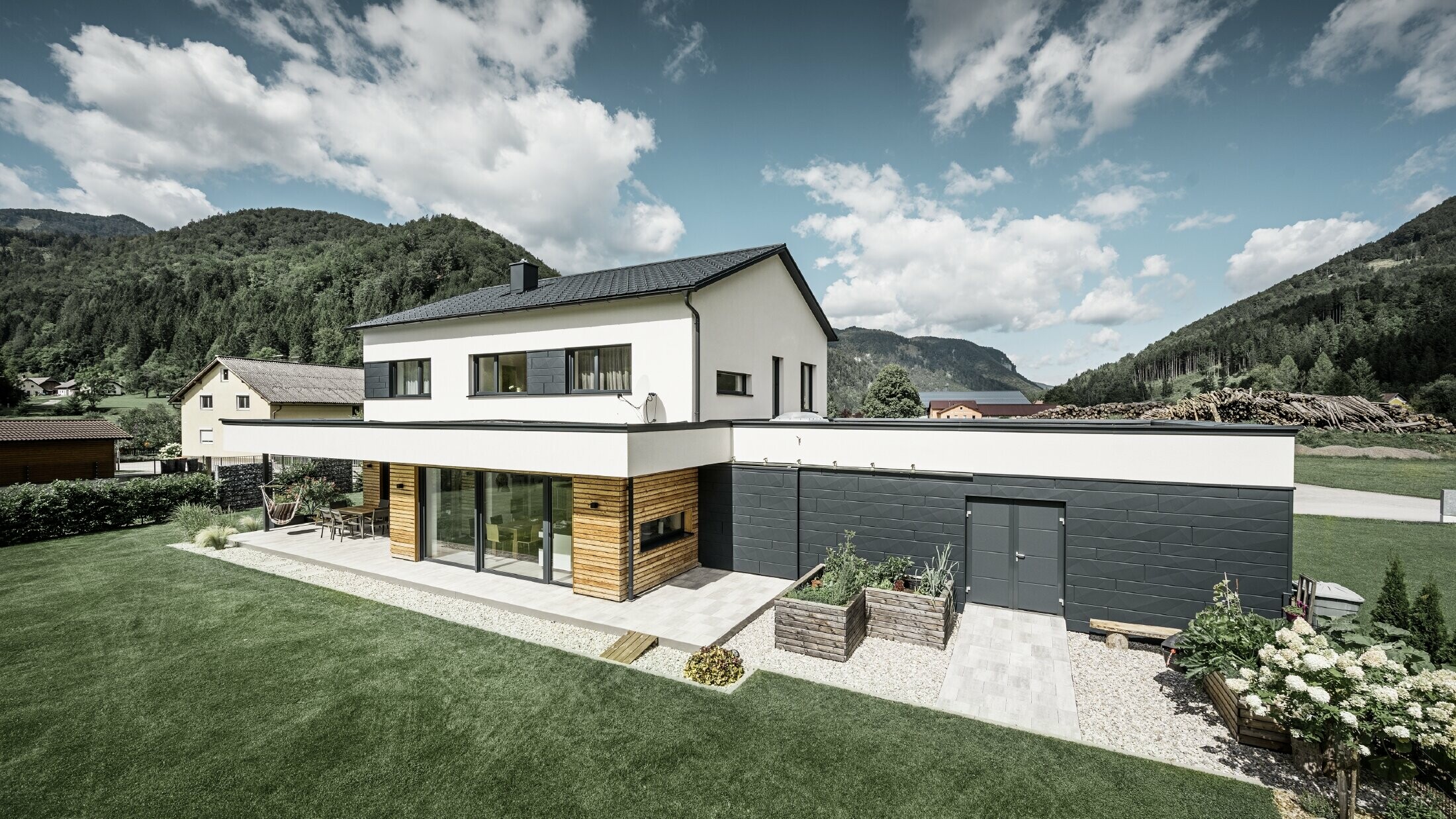 Moderne vrijstaande woning met wit gepleisterde gevel en PREFA Siding.X in P.10 antraciet, die een opvallend contrast vormt met de natuurlijke omgeving. Afgedekt met PREFA Dakpan, dakafvoer met PREFA hanggoot en regenpijp.