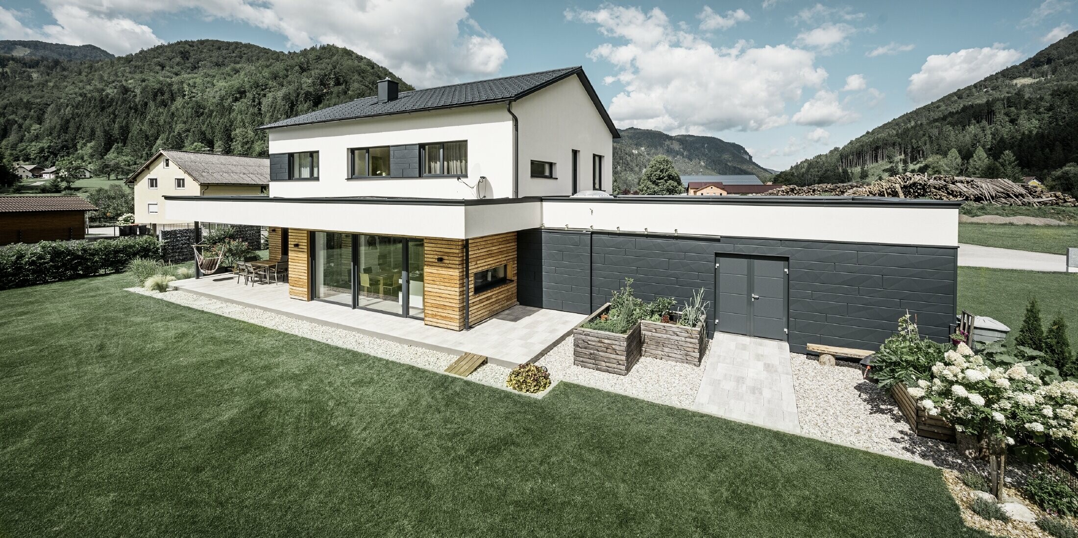 Moderne vrijstaande woning met wit gepleisterde gevel en PREFA Siding.X in P.10 antraciet, die een opvallend contrast vormt met de natuurlijke omgeving. Afgedekt met PREFA Dakpan, dakafvoer met PREFA hanggoot en regenpijp.