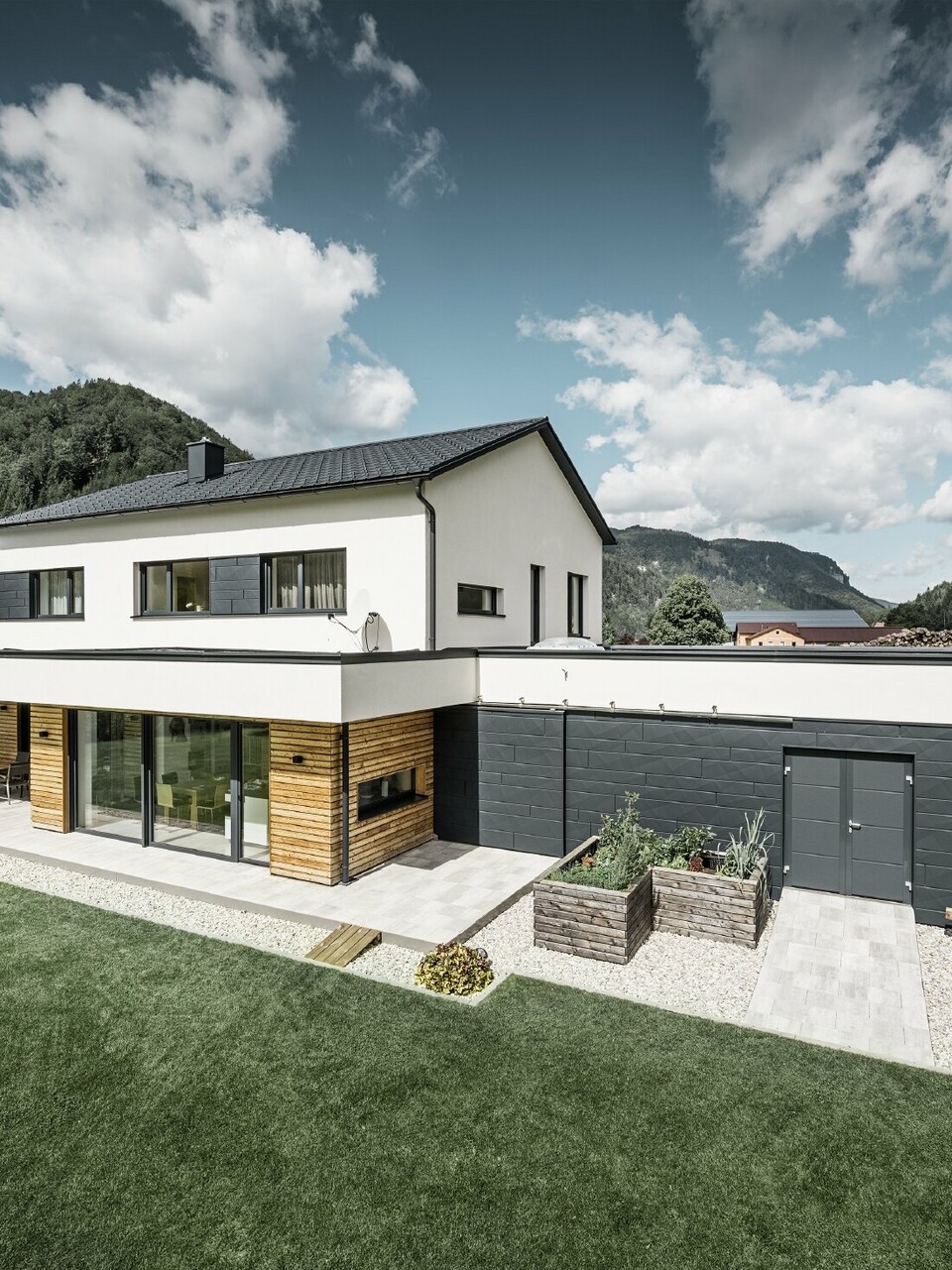Moderne vrijstaande woning met wit gepleisterde gevel en PREFA Siding.X in P.10 antraciet, die een opvallend contrast vormt met de natuurlijke omgeving. Afgedekt met PREFA Dakpan, dakafvoer met PREFA hanggoot en regenpijp.
