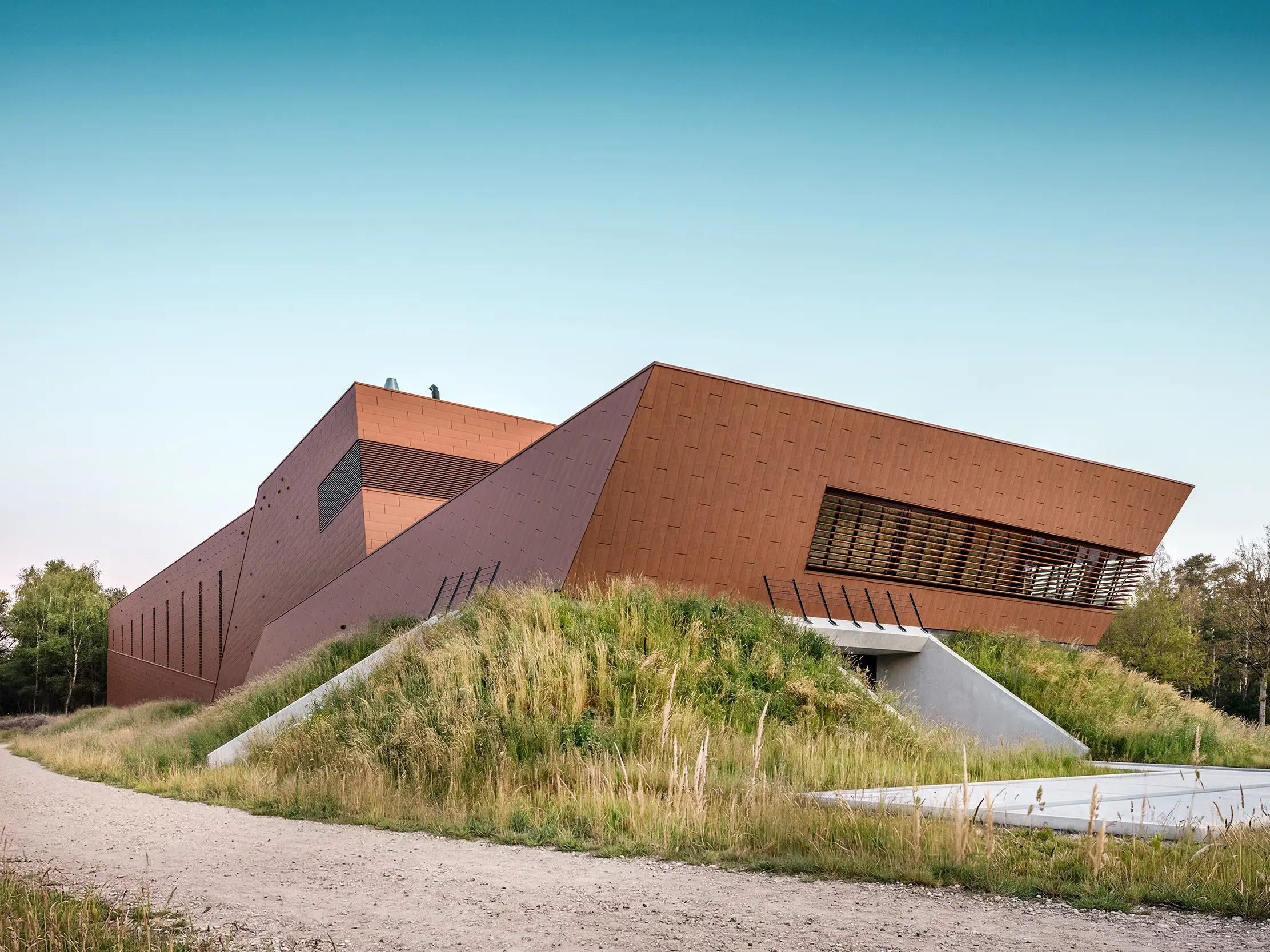 Architektonisch expressives Trinkwasserpumpwerk WML in Lottum mit skulptural gefalteten Baukörpern, eingebettet in eine naturnahe Böschungslandschaft. Die markante PREFA Siding.X Aluminiumfassade in warmer Rostoptik zieht mit ihrer facettierten, lebendigen Oberfläche alle Blicke auf sich und spielt eindrucksvoll mit Licht und Schatten. Horizontale Lamellen und präzise Linienführung betonen die dynamische Geometrie. Die leichten, langlebigen und vollständig recycelbaren PREFA Aluminium-Paneele stehen für höchste Witterungsbeständigkeit, nachhaltige Bauweise und eine wartungsarme Gebäudehülle.