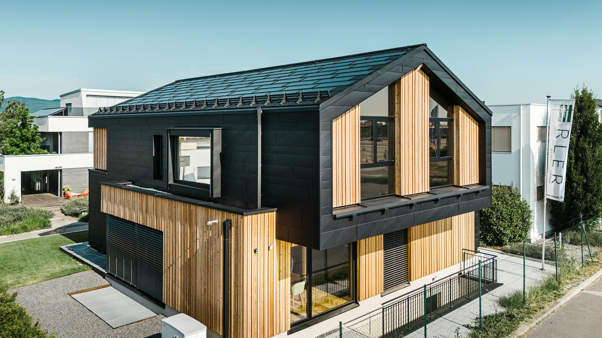 Voorzijde van het modelhuis Autark in het prefabhuiscentrum Blaue Lagune, Vösendorf (AT). Het dak is bedekt met zwarte PREFA solar-dakpan en dakpan R.16 met een hoogwaardige P.10-coating. De gevel bestaat uit een combinatie van verticale houten elementen en PREFA Siding.X in P.10 zwart, wat het gebouw een modern en duurzaam karakter geeft. Aan de rechterkant van de foto wappert de vlag van Erler Bau GmbH.