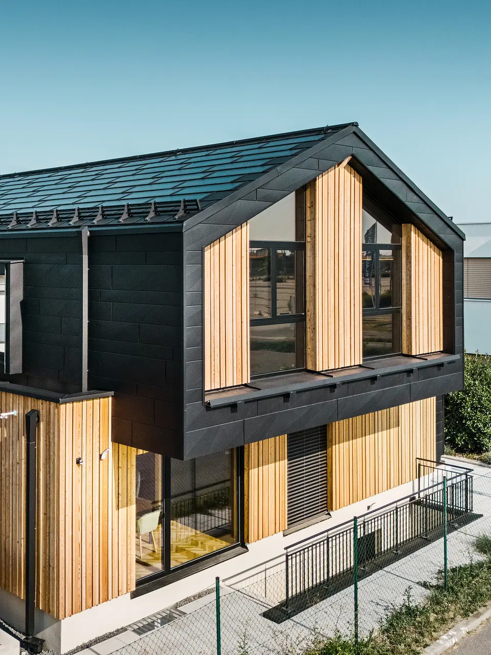 Voorzijde van het modelhuis Autark in het prefabhuiscentrum Blaue Lagune, Vösendorf (AT). Het dak is bedekt met zwarte PREFA solar-dakpan en dakpan R.16 met een hoogwaardige P.10-coating. De gevel bestaat uit een combinatie van verticale houten elementen en PREFA Siding.X in P.10 zwart, wat het gebouw een modern en duurzaam karakter geeft. Aan de rechterkant van de foto wappert de vlag van Erler Bau GmbH.