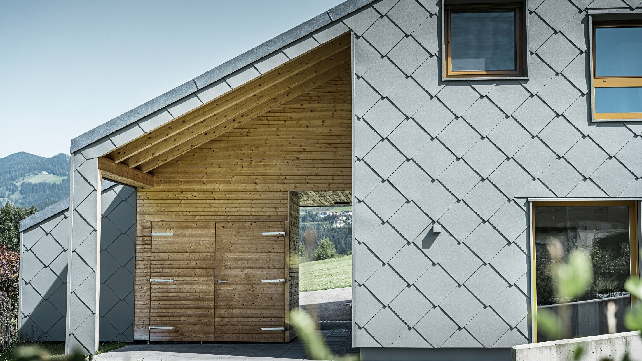 De overdekte toegangszone is voorzien van een horizontale houten bekleding; de rest van de gevel is bekleed met de grote PREFA gevellosange uit aluminium in lichtgrijs.