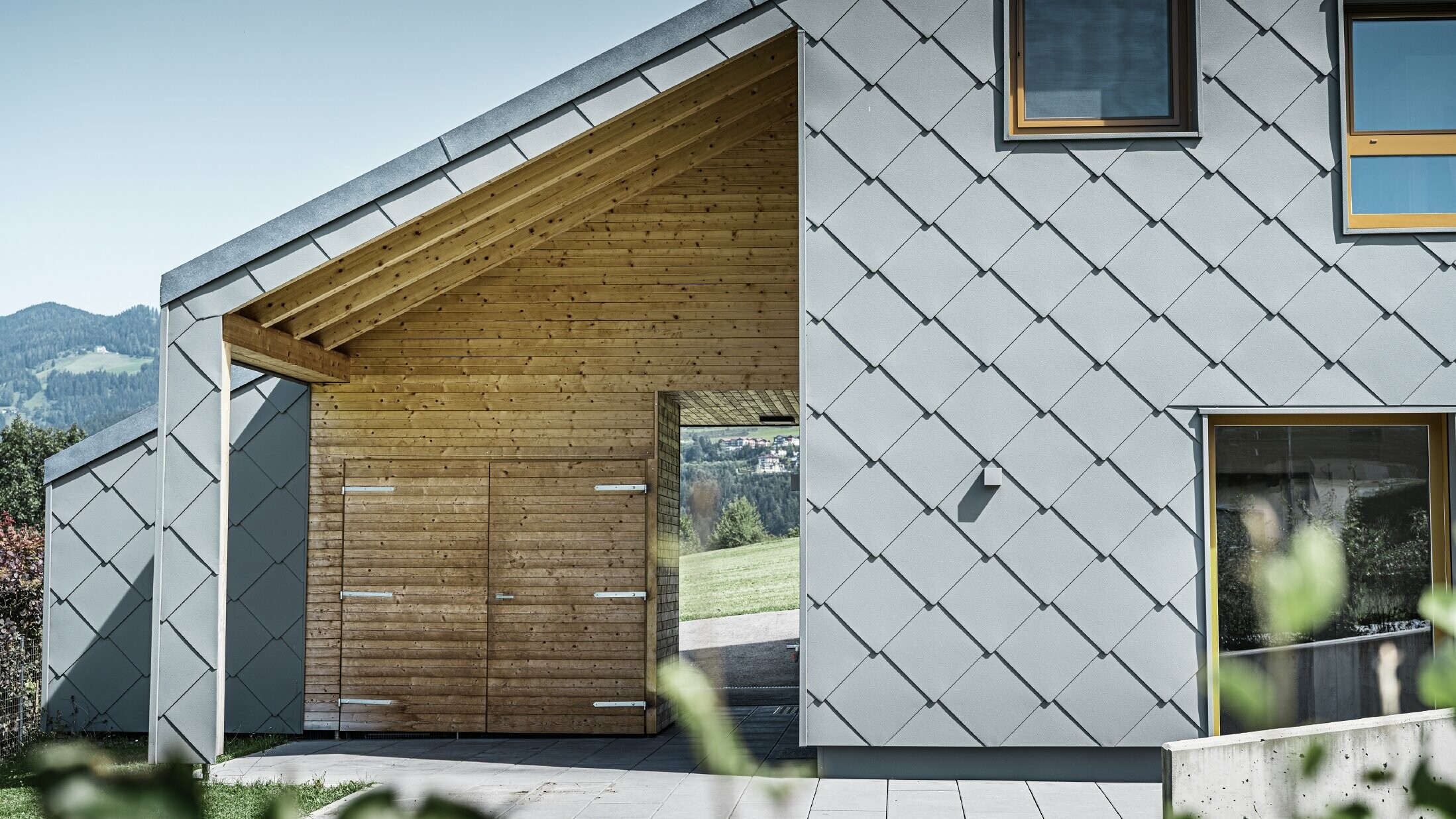 De overdekte toegangszone is voorzien van een horizontale houten bekleding; de rest van de gevel is bekleed met de grote PREFA gevellosange uit aluminium in lichtgrijs.