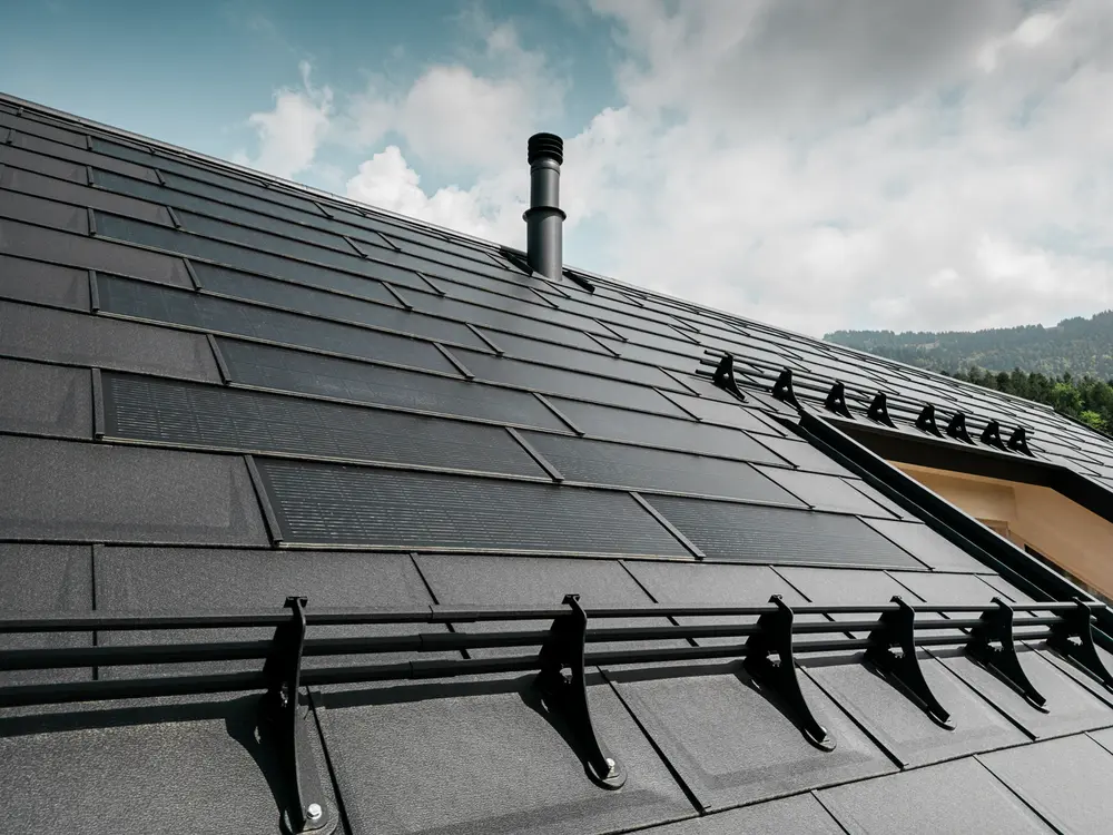Close-up van een modern dak van een huis, bedekt met zwarte PREFA solar dakpanelen en R.16 dakpanelen. Sneeuwschermen benadrukken de functionaliteit van het dak. Een dakraam en een schoorsteen onderbreken de uniformiteit van het dakoppervlak, dat zich uitstrekt tegen een achtergrond van beboste heuvels en een lichtblauwe lucht met weinig wolken.