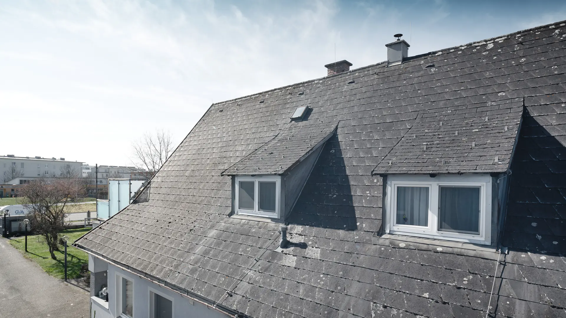 Detailfoto van een oud Eternit-dak met zichtbare slijtage en korstmos. Het dak heeft twee dakkapellen die zichtbaar zijn. Foto genomen vóór de dakrenovatie.