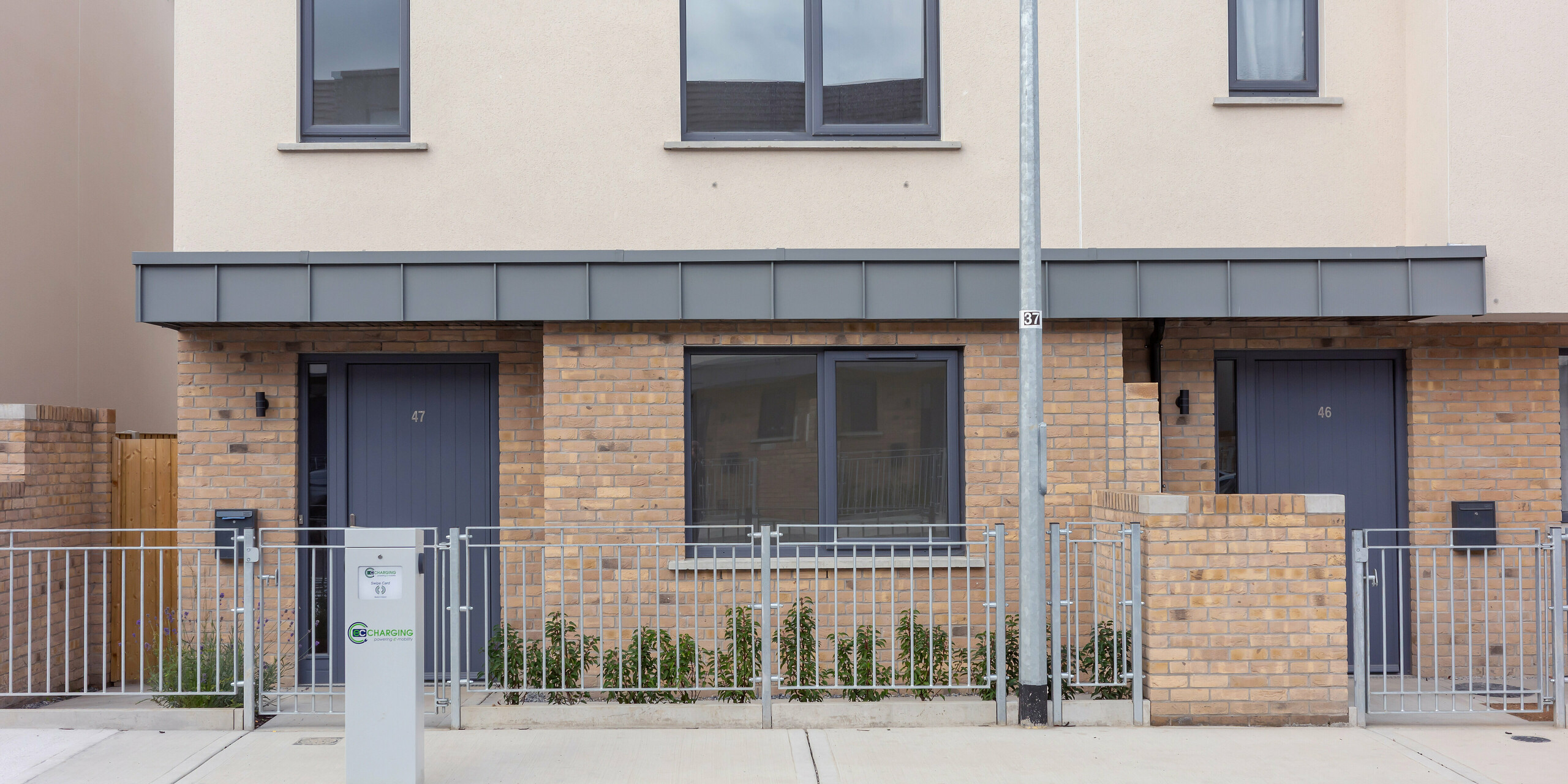 Das Bild zeigt den Eingangsbereich einer modernen Reihenhausanlage in Churchfields, geprägt von klaren Linien und harmonischen Farbflächen. Besonders ins Auge fällt die elegante Überdachung über den Haustüren, die mit PREFALZ in P.10 Hellgrau verkleidet ist. Die robuste Aluminiumoberfläche setzt einen modernen Akzent über dem warmen Backstein und ist nahezu wartungsfrei – ideal für große Wohnprojekte. Die einheitliche Fassadengestaltung und der gepflegte Vorgarten verleihen der Szene eine ruhige Ausstrahlung.