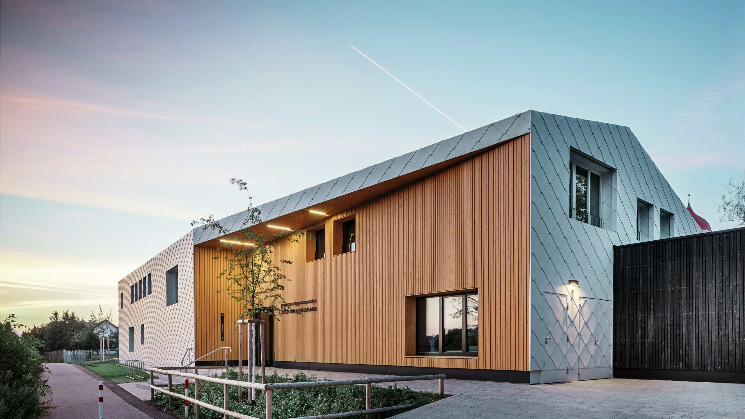 Foto genomen in de schemering van een modern kinderdagverblijf in Ulm. Het dak is ontworpen zonder oversteek. Het grootste deel van de gevel is bekleed met grote witte aluminium losanges van PREFA en de entree is uitgebreid bekleed met verticale houten panelen.