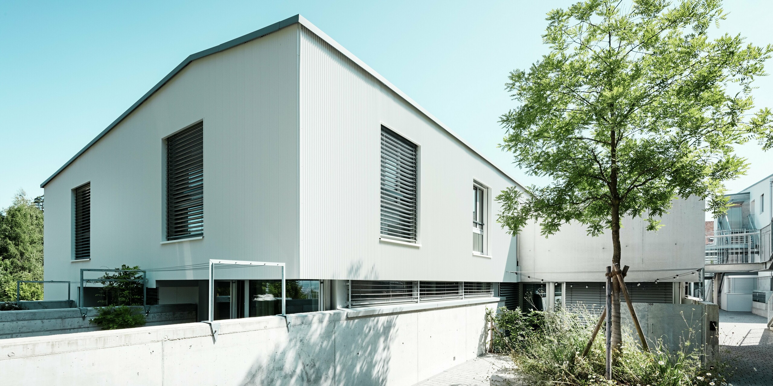 Moderne Tiny Homes am Zollikerberg in der Schweiz mit PREFALZ Dach in Patinagrau und Fassade aus PREFA Zackenprofil in P.10 Reinweiß. Die klar strukturierte Architektur wird durch horizontale Fensterbänder, Raffstores und Sichtbetonelemente ergänzt. Das hochwertige Aluminium überzeugt mit Witterungsbeständigkeit, geringem Wartungsaufwand und einem eleganten, zeitlosen Erscheinungsbild. Die Bäume in der Wohnanlage schaffen eine entspannte Wohnatmosphäre.