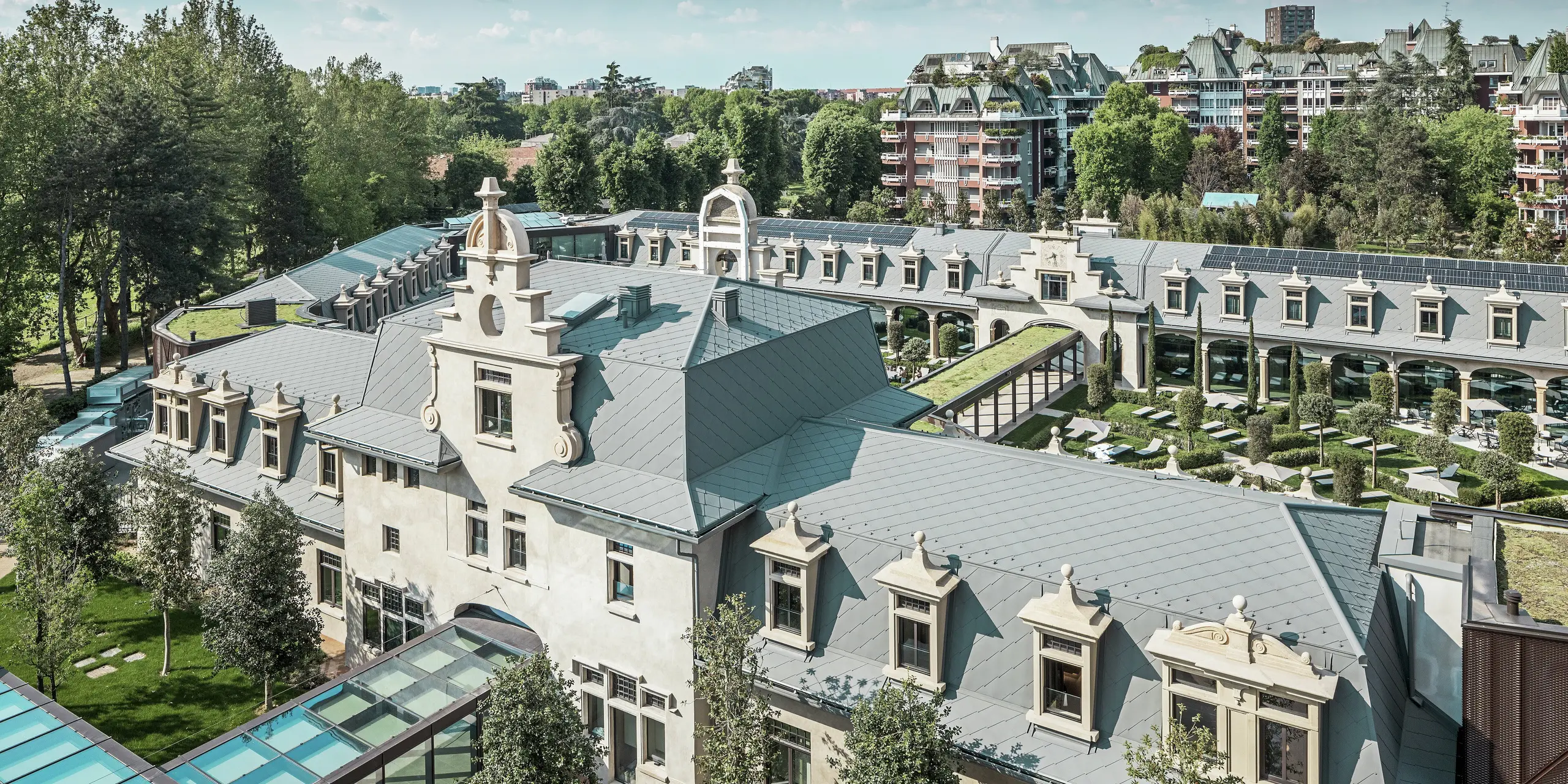 Totaalbeeld van het imposante complex “De Montel – Terme Milano” met monumentale art nouveau-architectuur en een moderne wellness-sfeer. De grootschalige dakbedekking met PREFA daklosange 44 × 44 in P.10 lichtgrijs geeft het complex een elegante uniformiteit. Geïntegreerde PV-modules, groene daken en glazen verbindingen tussen de gebouwen weerspiegelen de behoefte aan duurzaamheid en eigentijds design in een stedelijke context.
