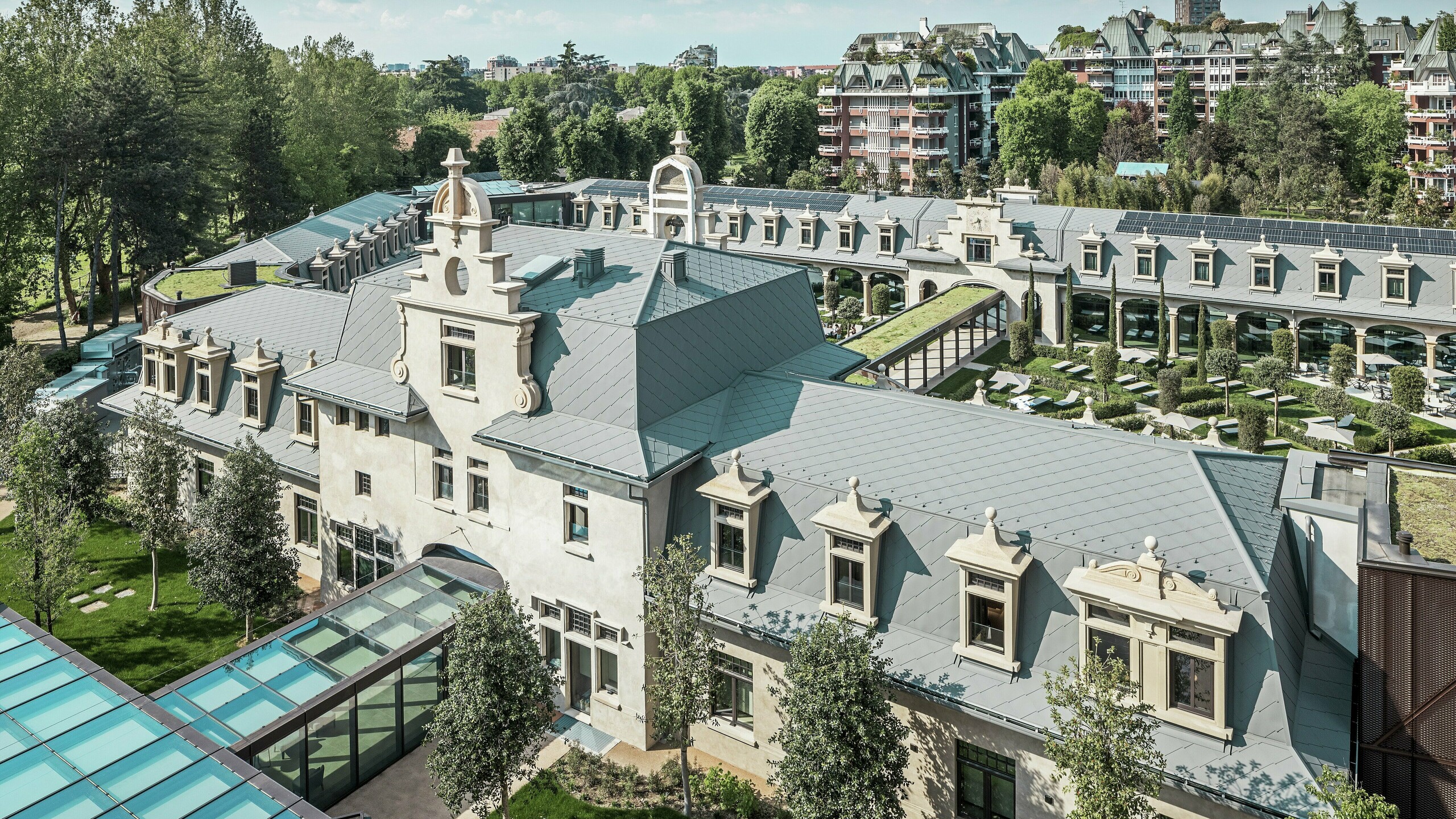 Totaalbeeld van het imposante complex “De Montel – Terme Milano” met monumentale art nouveau-architectuur en een moderne wellness-sfeer. De grootschalige dakbedekking met PREFA daklosange 44 × 44 in P.10 lichtgrijs geeft het complex een elegante uniformiteit. Geïntegreerde PV-modules, groene daken en glazen verbindingen tussen de gebouwen weerspiegelen de behoefte aan duurzaamheid en eigentijds design in een stedelijke context.