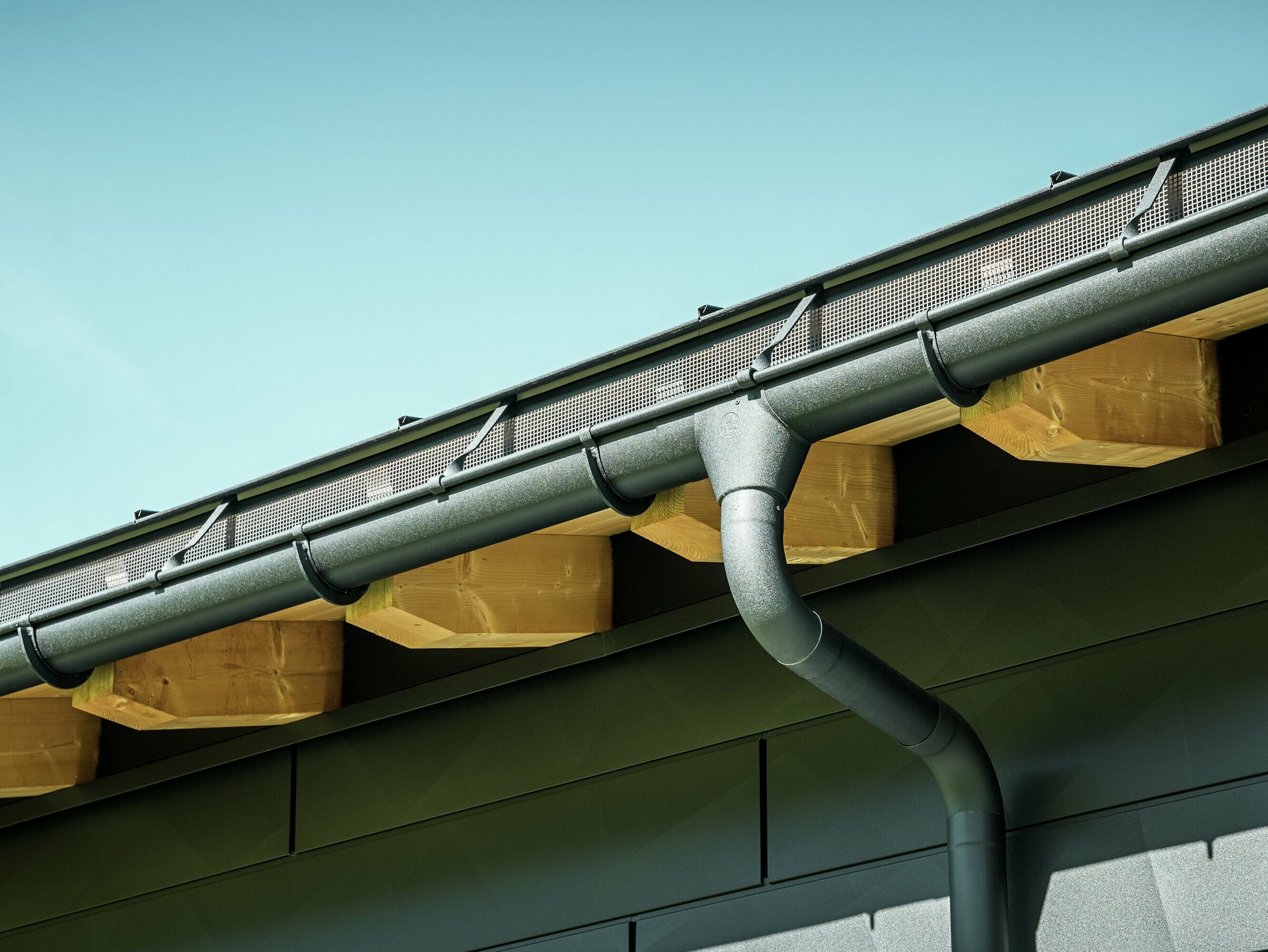 Close-up van de gevel van een moderne eengezinswoning in Ascharina, Zwitserland. De zijkanten van het gebouw zijn bekleed met de opvallende PREFA Siding.X gevelpanelen in P.10 zwart. De PREFA dakafvoersystemen met mastgoot en afvoerbuis zijn qua kleur op elkaar afgestemd.