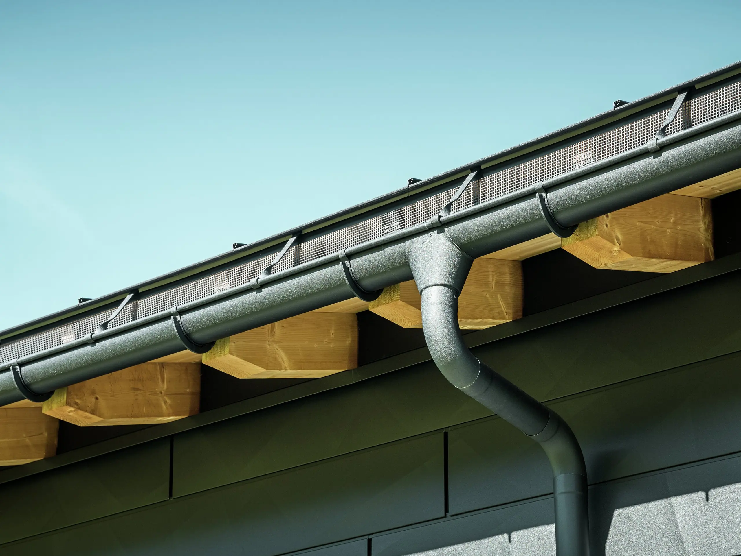 Close-up van de gevel van een moderne eengezinswoning in Ascharina, Zwitserland. De zijkanten van het gebouw zijn bekleed met de opvallende PREFA Siding.X gevelpanelen in P.10 zwart. De PREFA dakafvoersystemen met mastgoot en afvoerbuis zijn qua kleur op elkaar afgestemd.