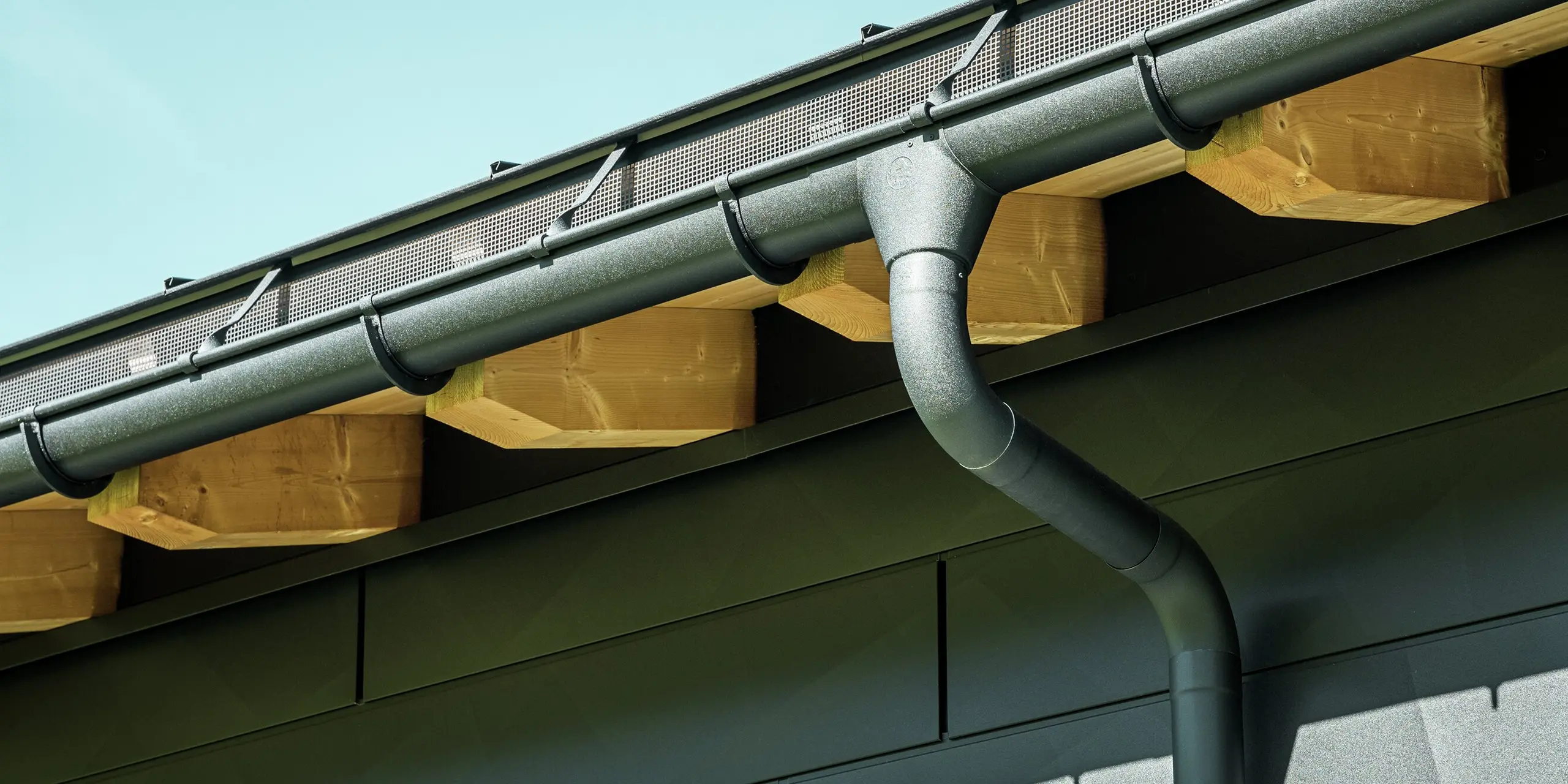 Close-up van de gevel van een moderne eengezinswoning in Ascharina, Zwitserland. De zijkanten van het gebouw zijn bekleed met de opvallende PREFA Siding.X gevelpanelen in P.10 zwart. De PREFA dakafvoersystemen met mastgoot en afvoerbuis zijn qua kleur op elkaar afgestemd.