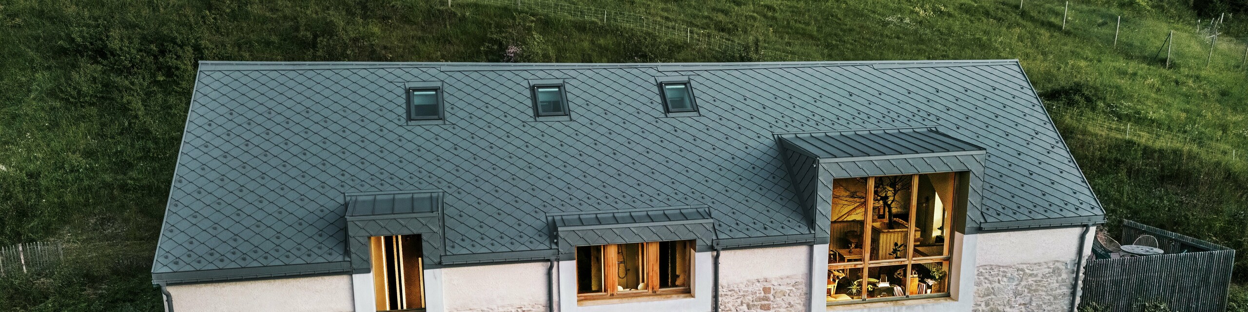 Abendansicht der restaurierten Scheune "La Grange à Gaby" in Engins, Frankreich. Das Gebäude kombiniert traditionelles Steinemauerwerk mit einem modernen PREFA Aluminiumdach aus 29 × 29 cm großen Rauten. Die großen Fenster und die beleuchteten Innenräume unterstreichen die einladende Atmosphäre des neuen kulturellen Zentrums.