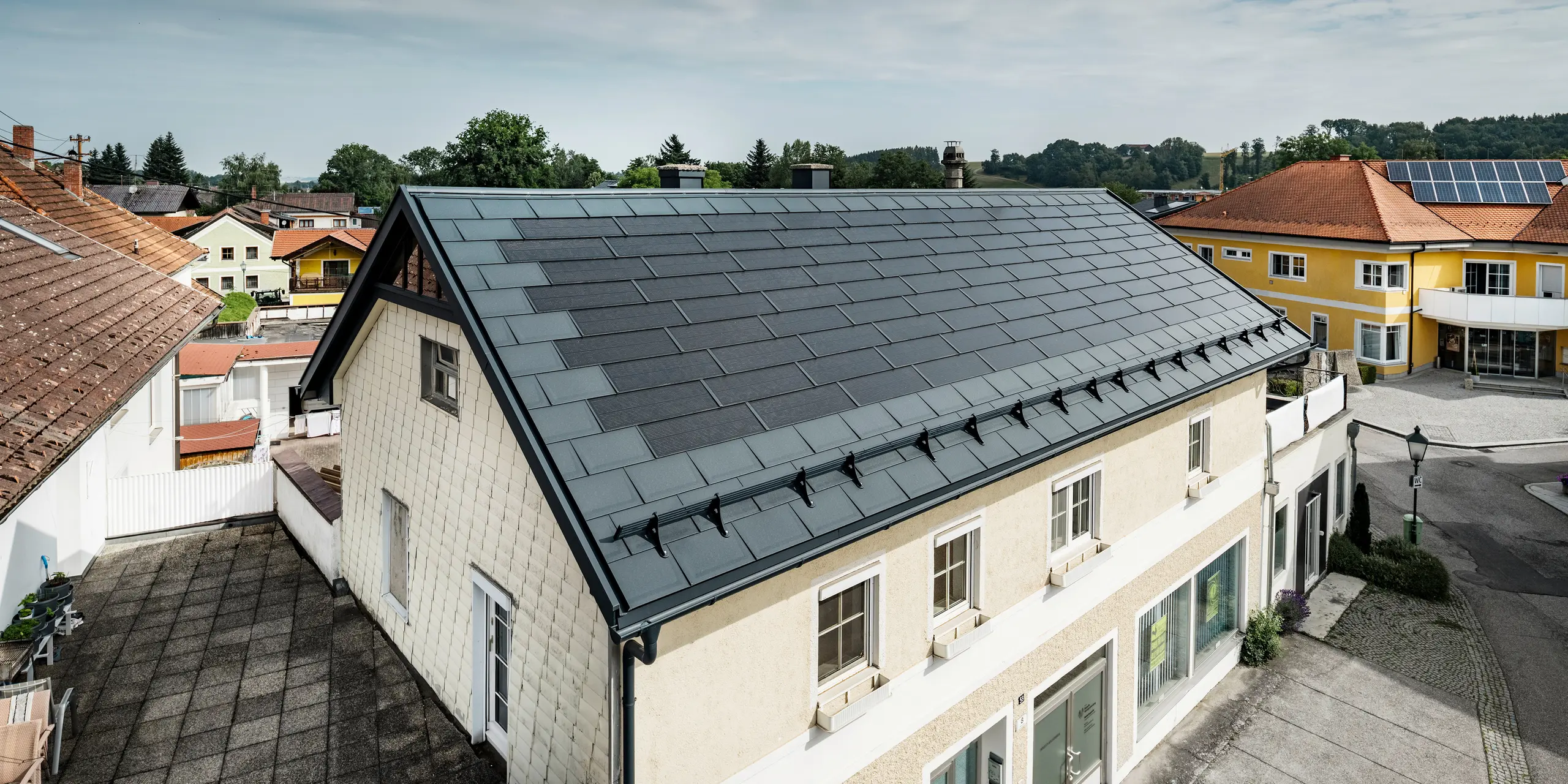 Bovenaanzicht van een vrijstaand huis in Mettmach,  Oostenrijk. Het oudere gebouw werd gerenoveerd met antracietkleurige PREFA-zonnedakplaten. De sneeuwremmers en de heldere lijnen van de zonnedakpanelen onderstrepen de strakke verwerking van de PREFA aluminium producten. Op de achtergrond contrasteert het oranjekleurige pannendak van een naburig gebouw. Op het dak van het naburige vrijstaande huis zijn fotovoltaïsche modules in een klassieke oplossing op het dak geïnstalleerd, die de veelzijdigheid van de energie-efficiënte dakconstructie demonstreren.