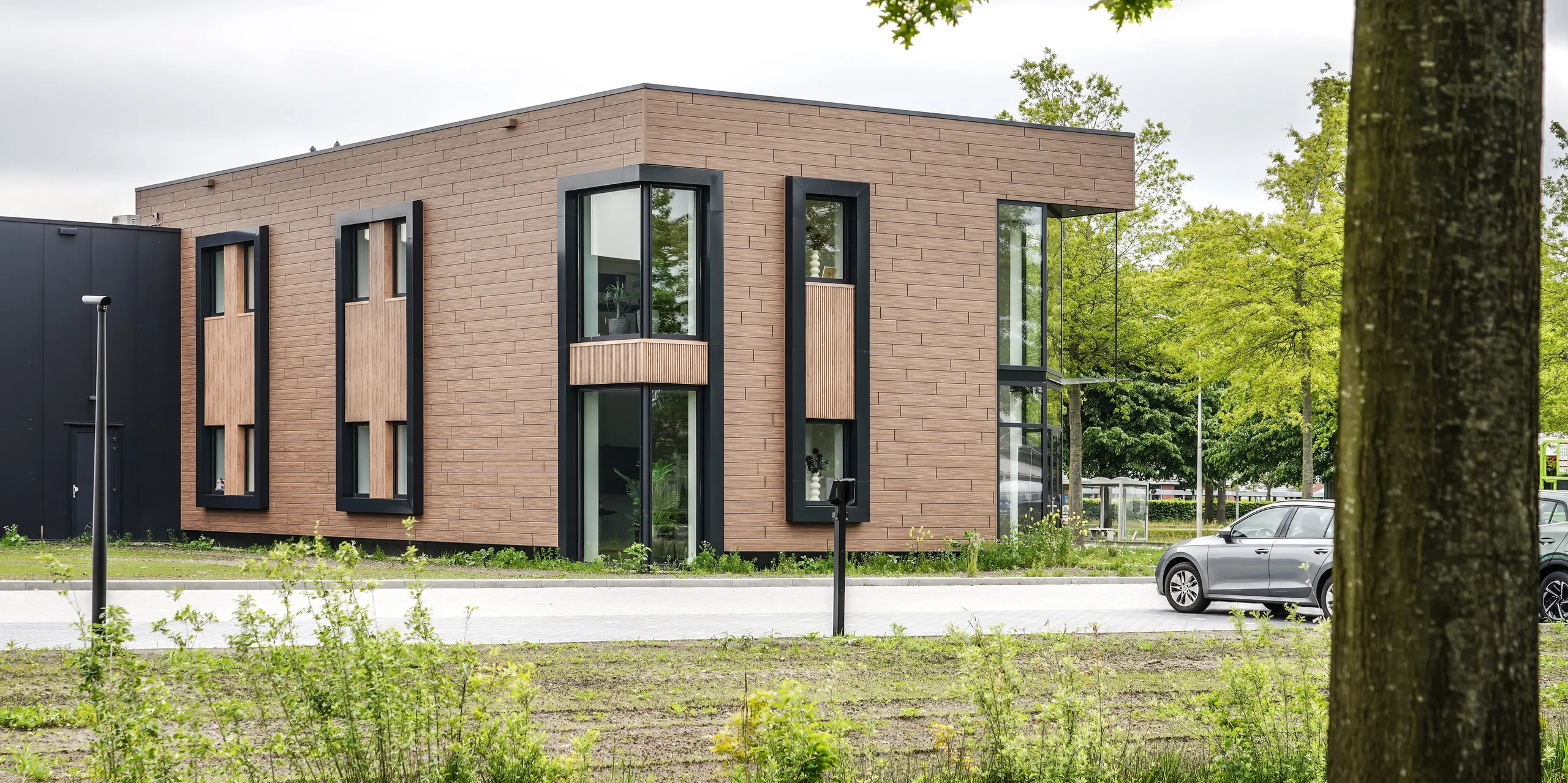Zwischen grünen Bäumen eingebettet präsentiert sich das Firmengebäude der Dokter Schoonmaakorganisatie in Wolvega als moderner Blickfang. Die walnussbraune PREFA Fassade mit Sidings in authentischer Holzoptik verleiht dem kubischen Baukörper eine warme Ausstrahlung. Schwarze Fensterrahmen, großzügige Verglasungen und markante Fassadenelemente schaffen ein spannendes Zusammenspiel aus Natur, Licht und zeitgenössischer Architektur.