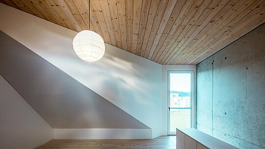 Das Bild zeigt einen minimalistischen Raum im Inneren des Einfamilienhauses in Freistadt. Die Holzdecke aus Weißtanne verleiht dem Raum eine warme und einladende Atmosphäre, die durch das schlichte Design und das natürliche Licht, das durch das Fenster auf der rechten Seite strömt, unterstützt wird. Das Fenster bietet einen Blick auf die grüne Umgebung und verstärkt das Gefühl von Offenheit. Der Raum ist von glatten, neutralen Wänden geprägt. Die Kombination aus Betonwand und Holzdecke schafft einen harmonischen Kontrast, der sowohl Eleganz als auch Natürlichkeit ausstrahlt. Die durchdachte Gestaltung des Innenraums spiegelt die Philosophie des gesamten Hauses wider: Funktionalität trifft auf ästhetische Klarheit und zeitlose Materialien.