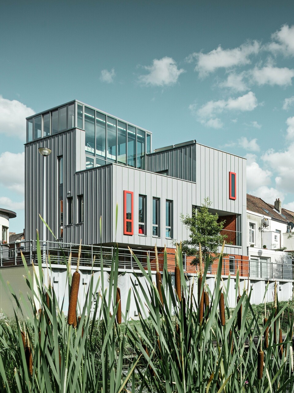 Architektonisches Highlight am Wasser: Das moderne Wohn- und Bürogebäude besticht durch eine PREFALZ Aluminiumfassade in Patinagrau mit vertikaler Stehfalzstruktur. Farblich abgesetzte Fenster in Rot setzen gezielte Akzente. Die Kombination aus Glas, Aluminium und rotem Klinker schafft ein harmonisches Gesamtbild – witterungsbeständig, pflegeleicht und langlebig.