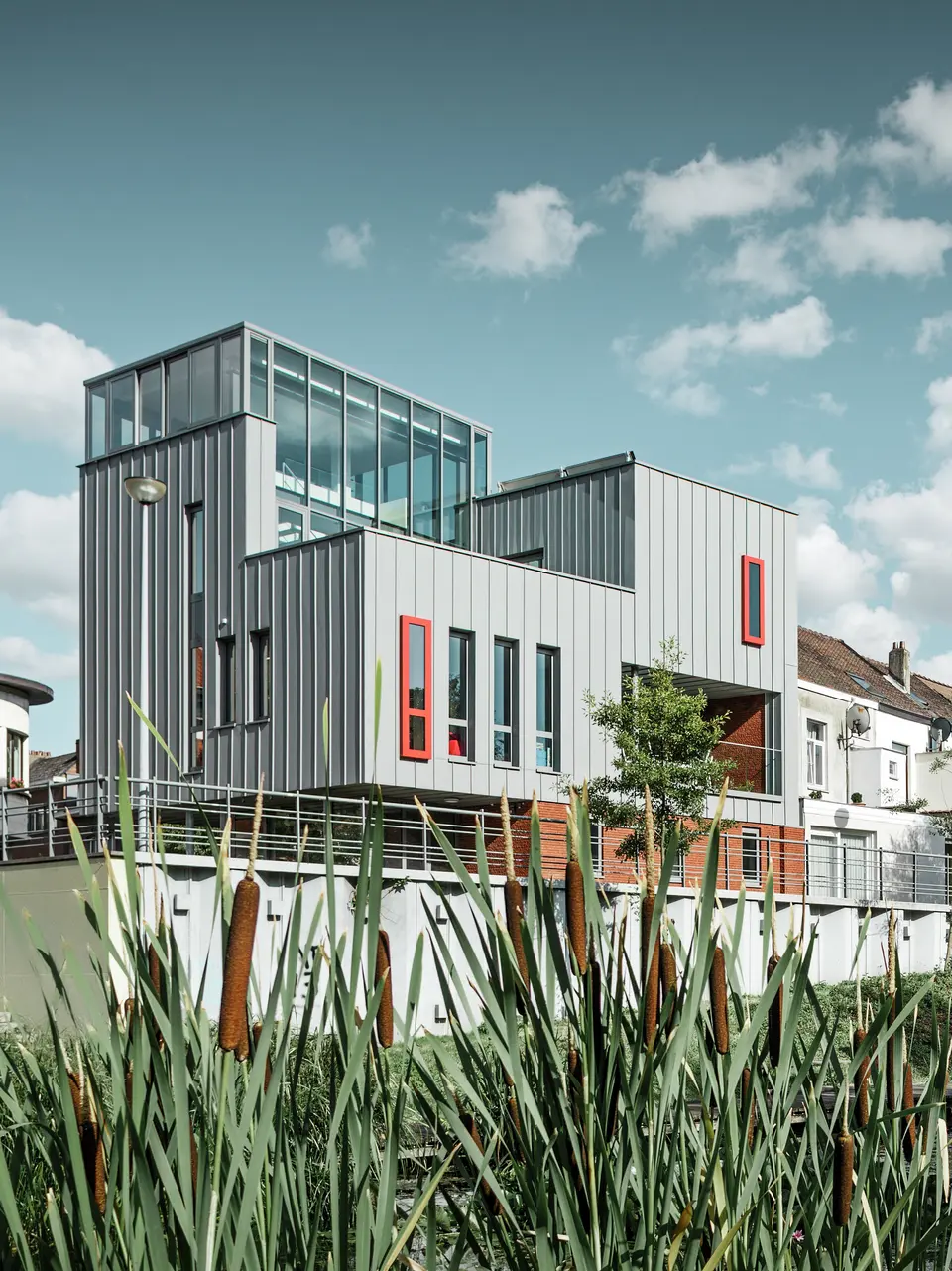 Architektonisches Highlight am Wasser: Das moderne Wohn- und Bürogebäude besticht durch eine PREFALZ Aluminiumfassade in Patinagrau mit vertikaler Stehfalzstruktur. Farblich abgesetzte Fenster in Rot setzen gezielte Akzente. Die Kombination aus Glas, Aluminium und rotem Klinker schafft ein harmonisches Gesamtbild – witterungsbeständig, pflegeleicht und langlebig.