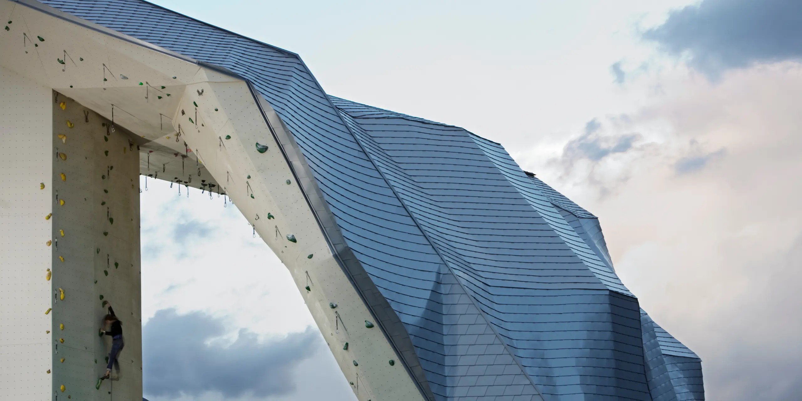 Im Vordergrund die Spitze des Kletterturms Imst mit Fassade bzw. Dach aus PREFA Dachschindeln in Silbermetallic. Am linken Bildrand eine Kletterin, am rechten Bildrand ein schöner Himmel in abendlicher Stimmung.