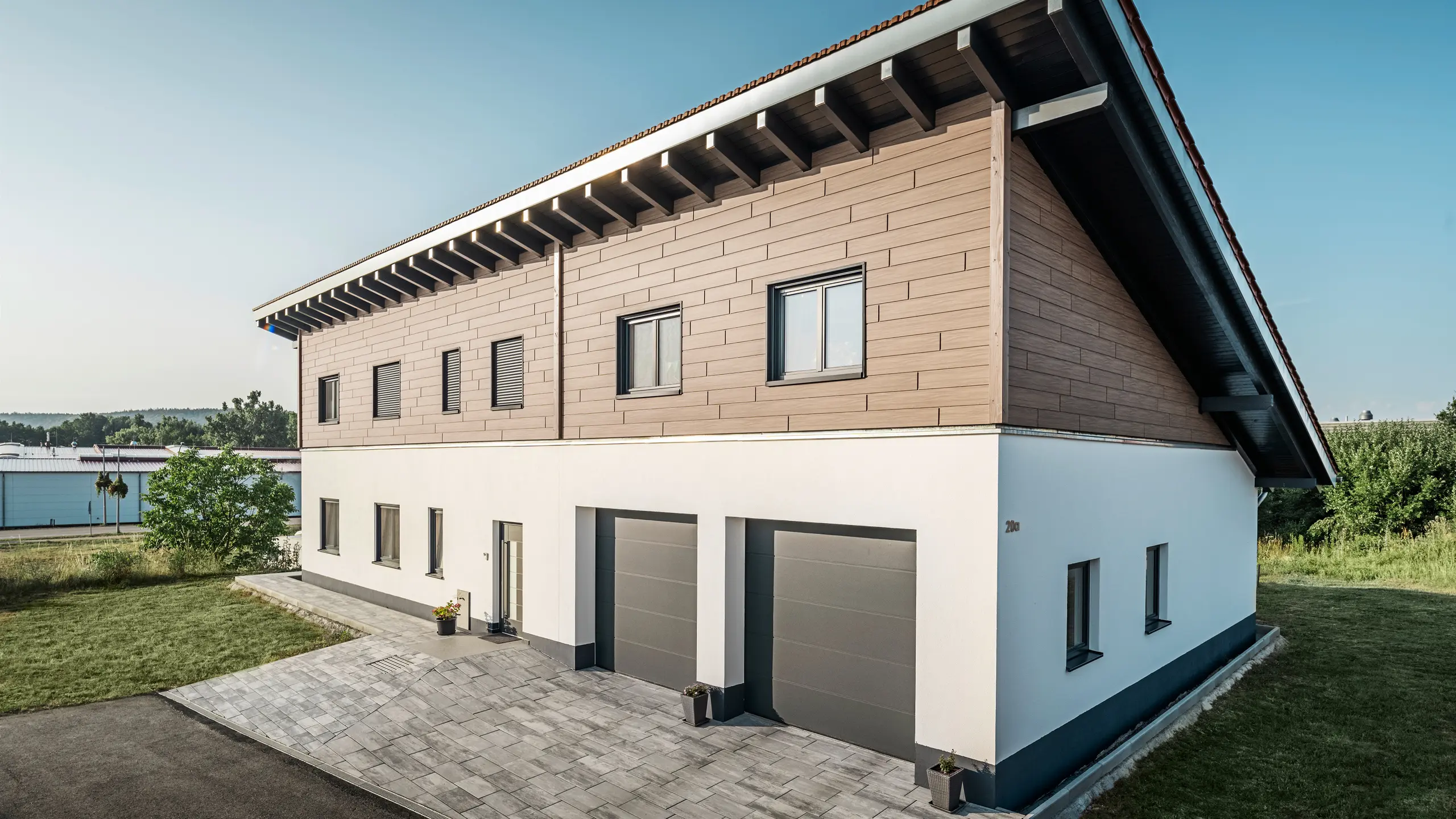 Zicht vanaf de zijkant op een vrijstaand huis met een lessenaarsdak in Bruck. De bovenste verdieping is bekleed met PREFA Sidings in walnoot Brown. Rechts op de foto zijn twee antracietkleurige garagedeuren te zien. De toegangsdeur bevindt zich in het midden van het gebouw. De plint is effen wit. 