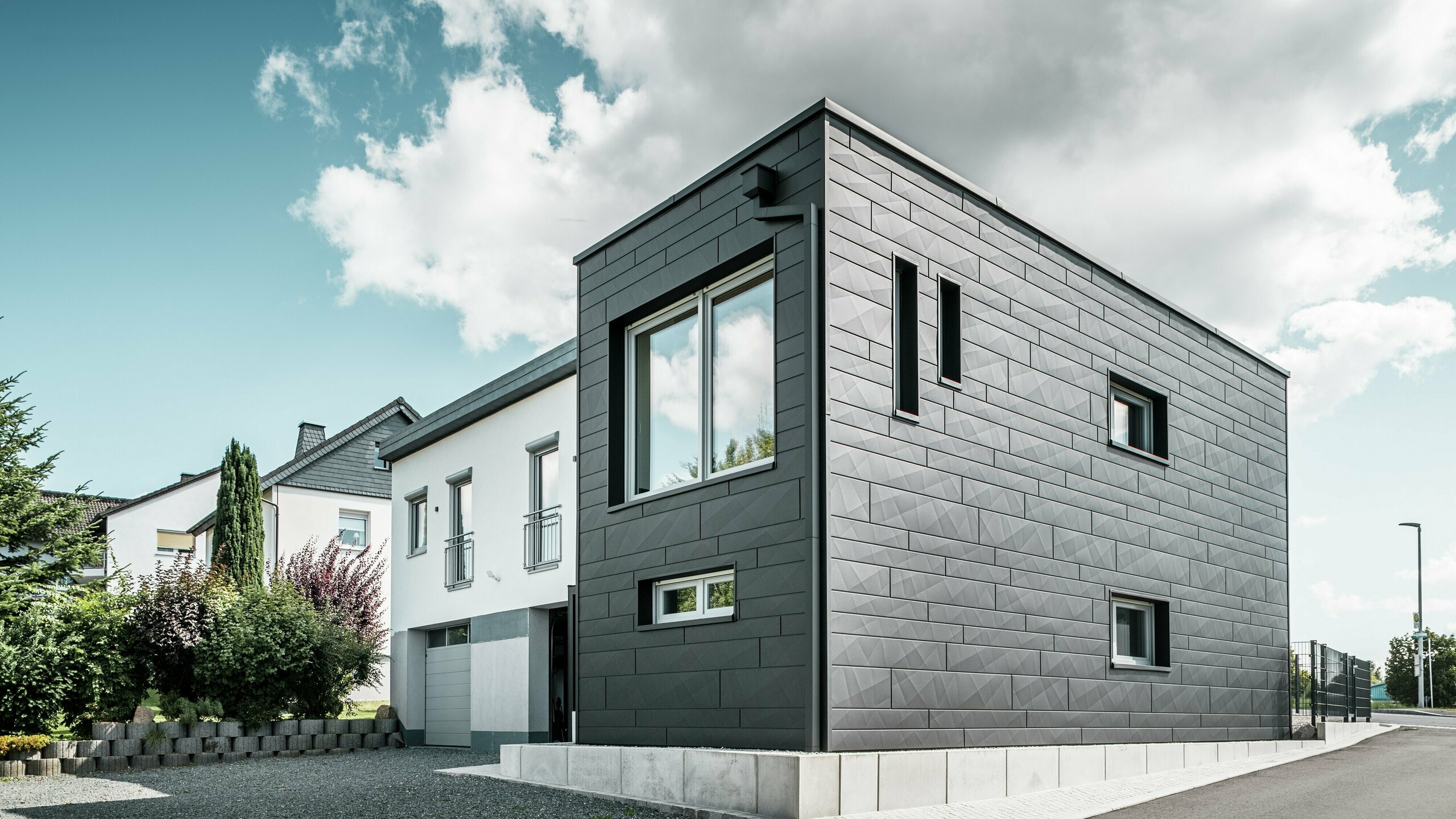 De stijlvolle en moderne gevel van de gerenoveerde eengezinswoning in Teuschnitz is voorzien van de duidelijk gestructureerde PREFA Siding.X-panelen in P.10 zwart. De geometrische opstelling van de hoogwaardige plaatpanelen geeft het gebouw een opvallende, tijdloze uitstraling. De ramen zijn omlijst met zwarte aluminium composietplaten, wat het ontwerp harmonieus afrondt. De geïntegreerde PREFA dakafvoersystemen met vierkante buis zorgen voor een functionele en esthetische oplossing.