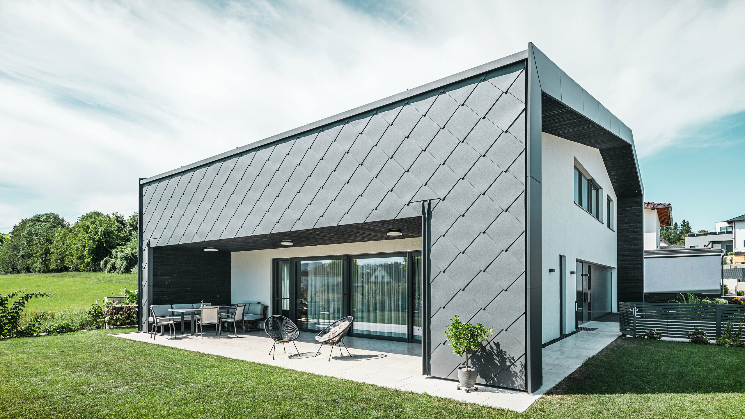 Verzorgde tuin van een eengezinswoning in Aschach. Het gebouw is stijlvol bekleed met donkergrijze PREFA aluminium gevellosange 44 × 44. Het overdekte terras biedt een gezellige zitplaats met uitzicht op het weelderige groene gazon. De elegante aluminium bekleding in combinatie met witte pleisterwerk zorgt voor een moderne en weerbestendige architectuur. Grote ramen laten veel daglicht binnen en creëren een open, lichte sfeer.