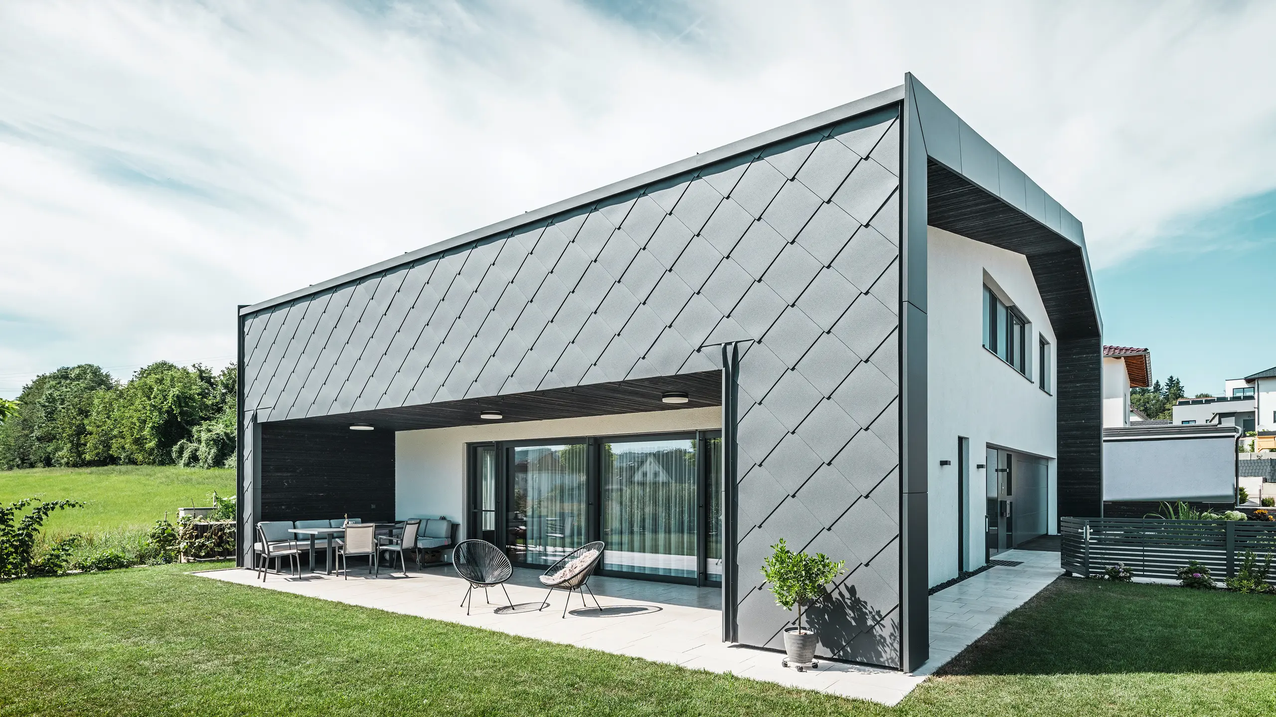Verzorgde tuin van een eengezinswoning in Aschach. Het gebouw is stijlvol bekleed met donkergrijze PREFA aluminium gevellosange 44 × 44. Het overdekte terras biedt een gezellige zitplaats met uitzicht op het weelderige groene gazon. De elegante aluminium bekleding in combinatie met witte pleisterwerk zorgt voor een moderne en weerbestendige architectuur. Grote ramen laten veel daglicht binnen en creëren een open, lichte sfeer.