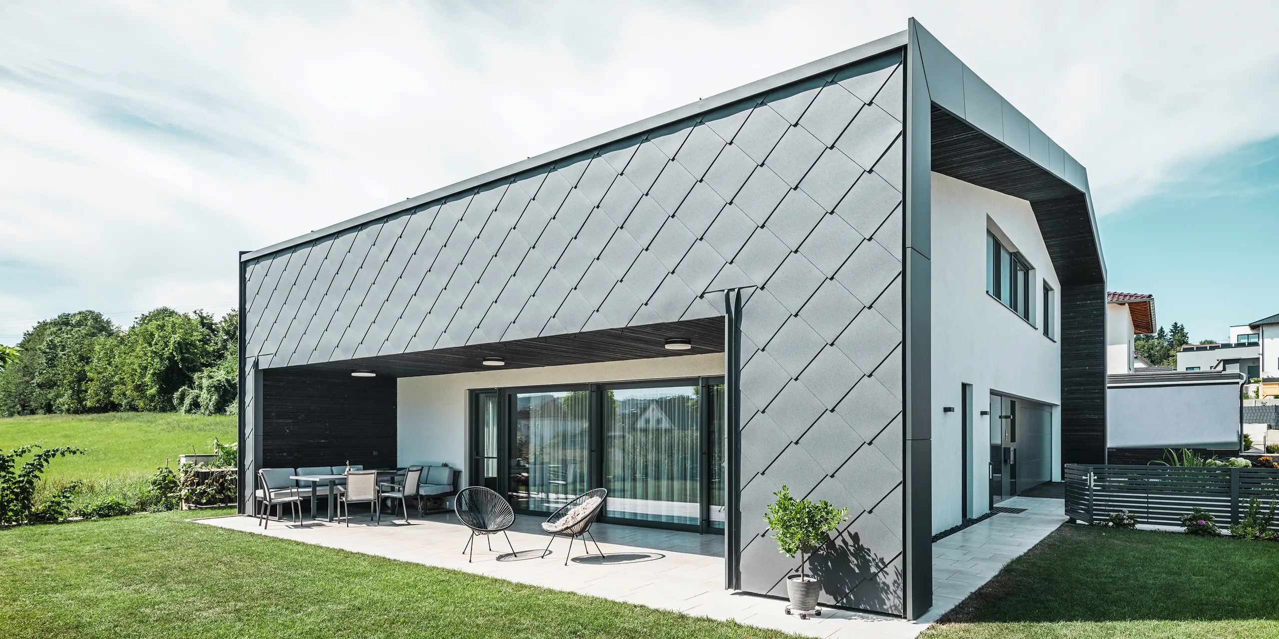 Verzorgde tuin van een eengezinswoning in Aschach. Het gebouw is stijlvol bekleed met donkergrijze PREFA aluminium gevellosange 44 × 44. Het overdekte terras biedt een gezellige zitplaats met uitzicht op het weelderige groene gazon. De elegante aluminium bekleding in combinatie met witte pleisterwerk zorgt voor een moderne en weerbestendige architectuur. Grote ramen laten veel daglicht binnen en creëren een open, lichte sfeer.