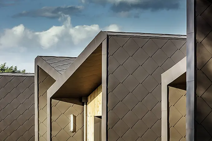 Das Bild zeigt eine moderne Fassade, verkleidet mit PREFA Dach- und Wandprofilen (44 × 44 mm) in Walnussbraun. Die Profilstruktur der Aluminiumverkleidung betont die klaren Linien und bietet eine robuste und langlebige Lösung für Dach- und Fassadensysteme. Die Kombination aus dunklen Metallprofilen und warmen Holzelementen verleiht dem Gebäude eine elegante und zeitlose Optik. Diese moderne und hochwertige Architektur eignet sich ideal für energieeffiziente und ästhetisch ansprechende Bauprojekte.