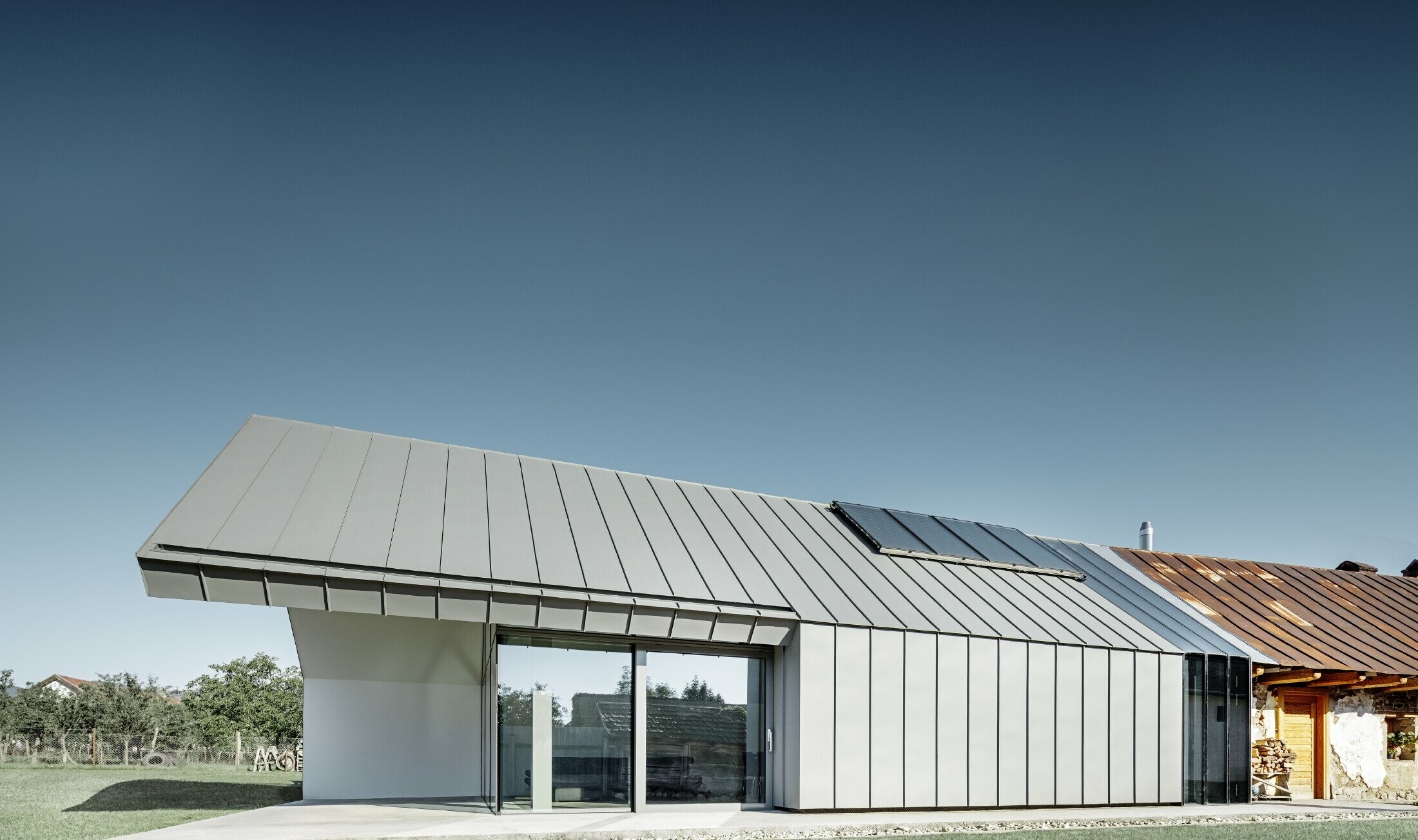 Modern architectenbureau/woning gebouwd tegen een oude boerderij, bekleed met staande naden en gebogen staande naden van PREFA in lichtgrijs