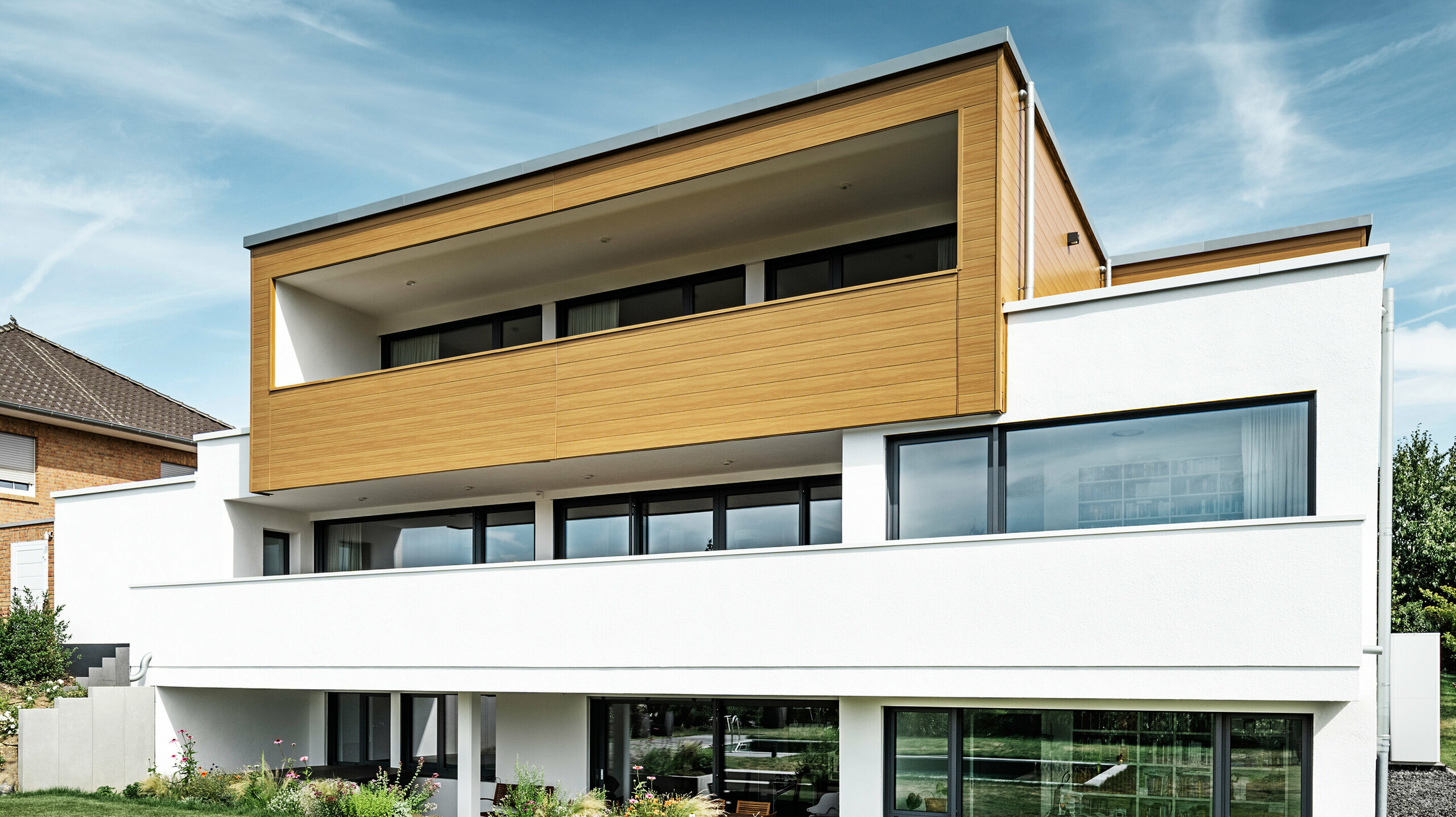 De achterkant van een modern vrijstaand huis in Usingen, Duitsland. Het groene gazon, een zwembad en een jonge tuinboom zijn onderaan de foto te zien. Het in het oog springende huis heeft een witte bouwbasis van een bepleisterde gevel, een aluminium gevel op de bovenverdieping van PREFA Sidings in eiken naturel en grote raamoppervlakken met een antracietkleurig kozijn. Van een afstand is het buurhuis met een bakstenen gevel aan de linkerkant van de foto te zien.  Vertaald met DeepL.com (gratis versie)